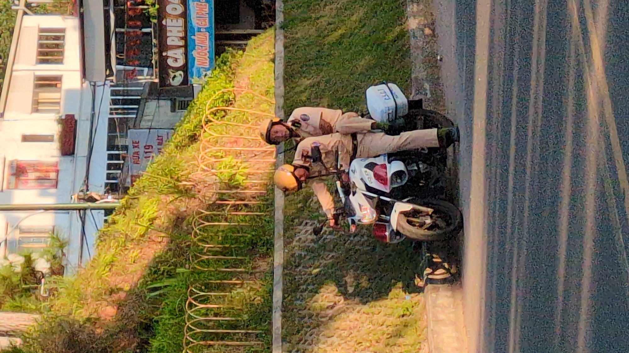 Two individuals, appearing to be police officers based on their uniforms and helmets, are seated on a white motorcycle. The officer in the front is operating the motorcycle, while the second officer is a passenger. They are positioned on a paved road next to a concrete wall with a metal fence. Behind them, a vegetated embankment rises, with buildings visible at the top. A sign with Vietnamese text, including "RUA XE 119" and "CA PHE O", is partially visible in the background. The scene suggests an urban or semi-urban environment in Da Lat, Vietnam, possibly on a street or hillside road, with commercial establishments in the background.