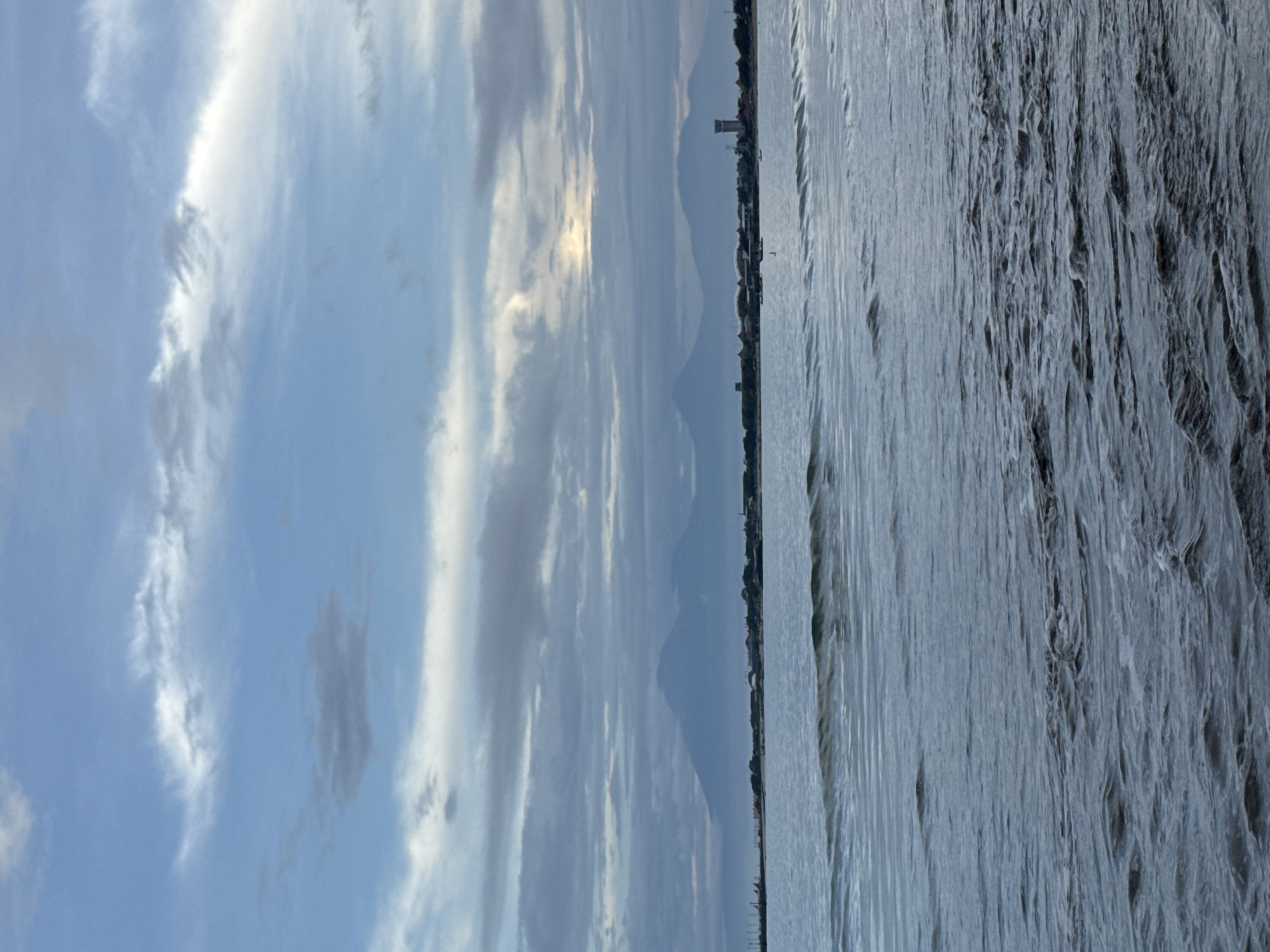 The image presents a landscape likely situated in Kuta Selatan, Indonesia, featuring a body of water in the foreground, potentially the sea or a large lake. The water surface displays subtle ripples and varying degrees of texture, suggesting slight wave activity.
In the mid-ground, a thin strip of land is visible, marked by what appears to be a coastline or the edge of the land mass, dotted with buildings and what may be vegetation. This indicates the presence of a settlement or urban area along the shoreline. The arrangement of structures and the presence of a taller tower-like structure suggest a built environment.
The background is dominated by a sky filled with a complex arrangement of clouds. The clouds vary in density and shape, with some areas appearing more illuminated, possibly by sunlight filtering through. The presence of mountains are visible in the distance.