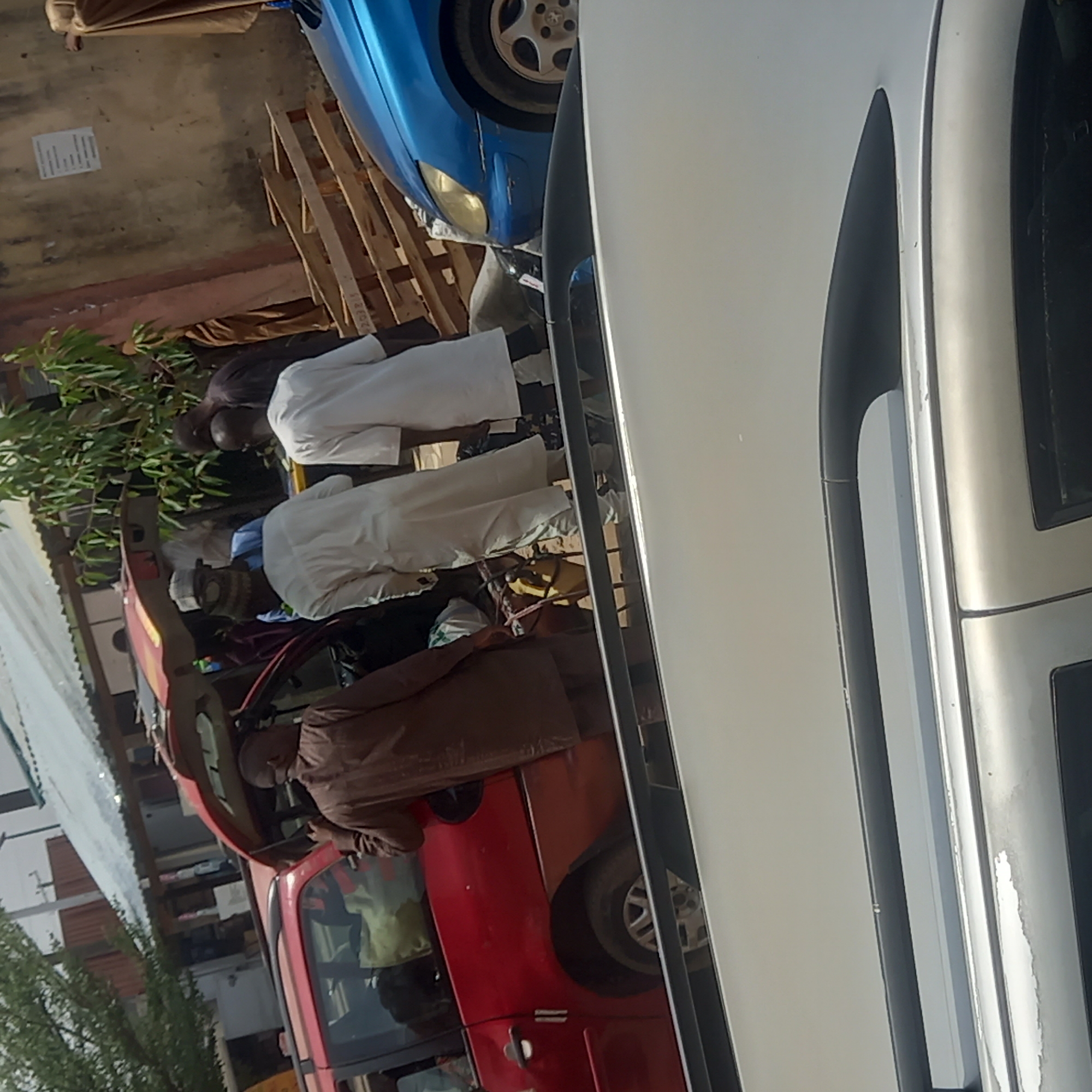 The image depicts an outdoor daytime scene in Dadi Riba, Nigeria, under clear skies. Several men are present, engaged around multiple vehicles. In the foreground on the right, a large portion of a silver or white car is visible. To the left, a blue car is partially seen, parked near stacked wooden pallets. A man in a brown garment stands next to the blue car. In the mid-ground, a red vehicle, possibly a small truck or van, has its door open. A man in a brown outfit is leaning into this red vehicle, appearing to attend to its interior. Two men wearing white traditional clothing are also visible near the red vehicle; one is bending forward, potentially in prayer or focused on an item on the ground.
Green foliage and distant structures are visible in the background. The ground is a mix of concrete and dirt. A white sheet of paper with printed text is on the ground near the blue car, but its content is illegible. On one of the wooden pallets, the numbers "2023" are partially discernible. The activity suggests a typical day in the area, possibly involving vehicle use, social interaction, or local commerce.