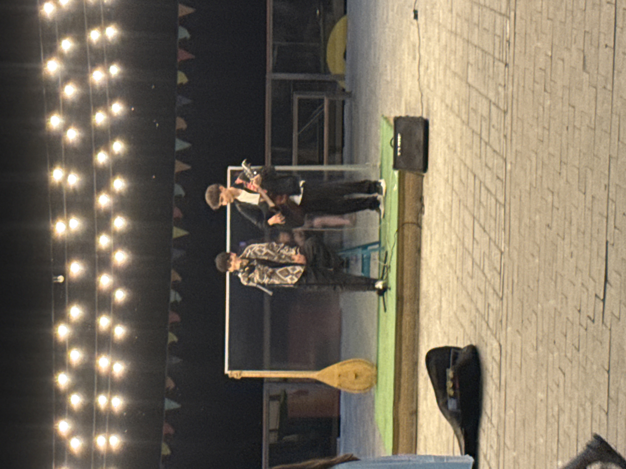 Two young men are performing on an outdoor stage under a string of lights in Pavlodar, Kazakhstan. The man on the left is seated on a blue stool, wearing a patterned jacket and black pants, and holding a microphone. To his right, a man stands playing an acoustic guitar. A long-necked string instrument, possibly a dutar, is propped up beside the stage. The stage itself is a raised platform covered in green material, with a small black amplifier positioned at its base. The ground around the stage is paved with light-colored bricks. In the foreground, a black instrument case lies on the pavement. The background consists of a dark wall and some fencing, with more string lights visible overhead.