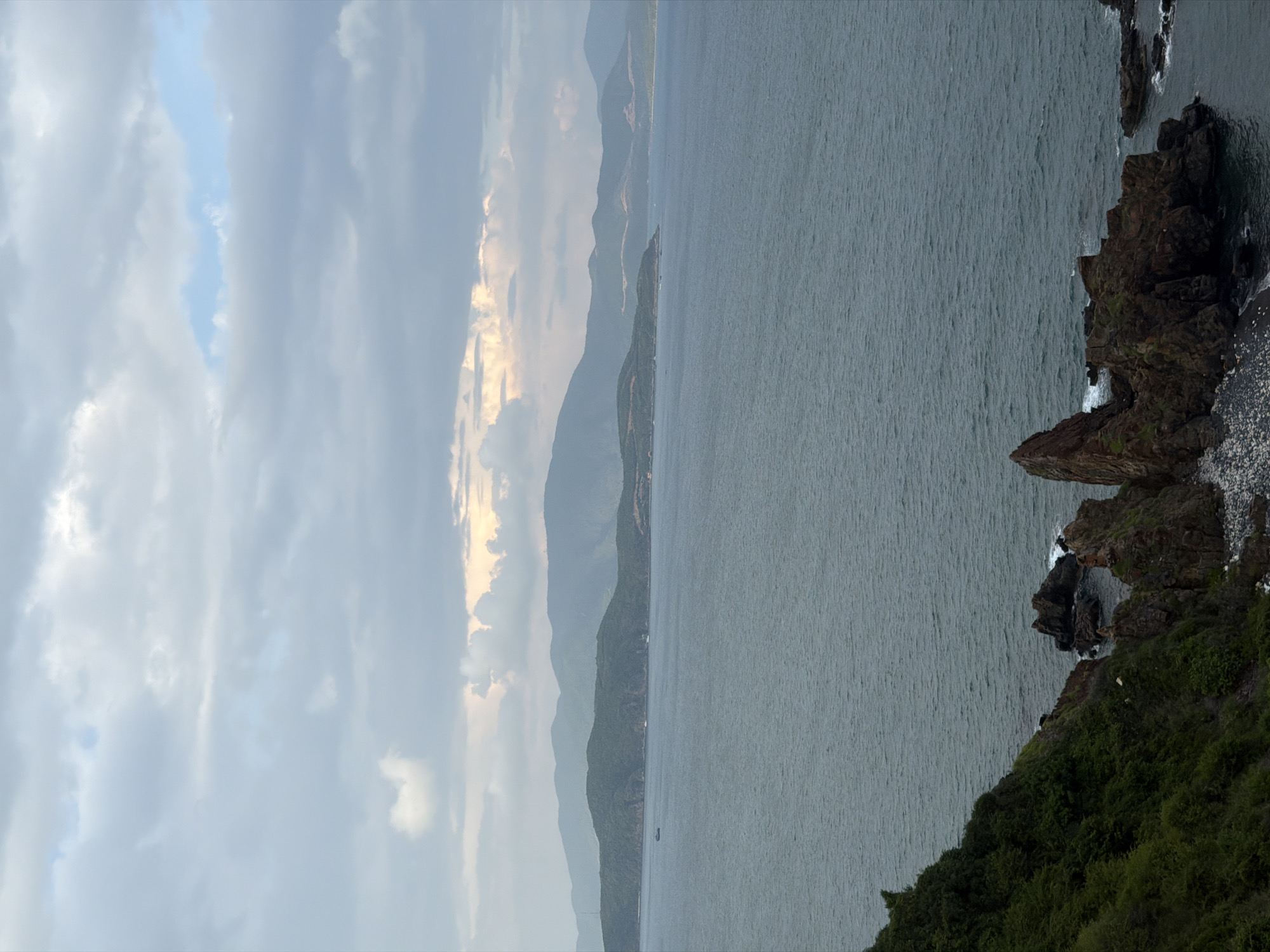 The image depicts a coastal scene in Nam Nha Trang Ward, Vietnam. It features the ocean meeting a rocky shoreline with green vegetation. Mountains are visible in the background. The sky is cloudy, with some light breaking through. A small boat is visible on the water.
