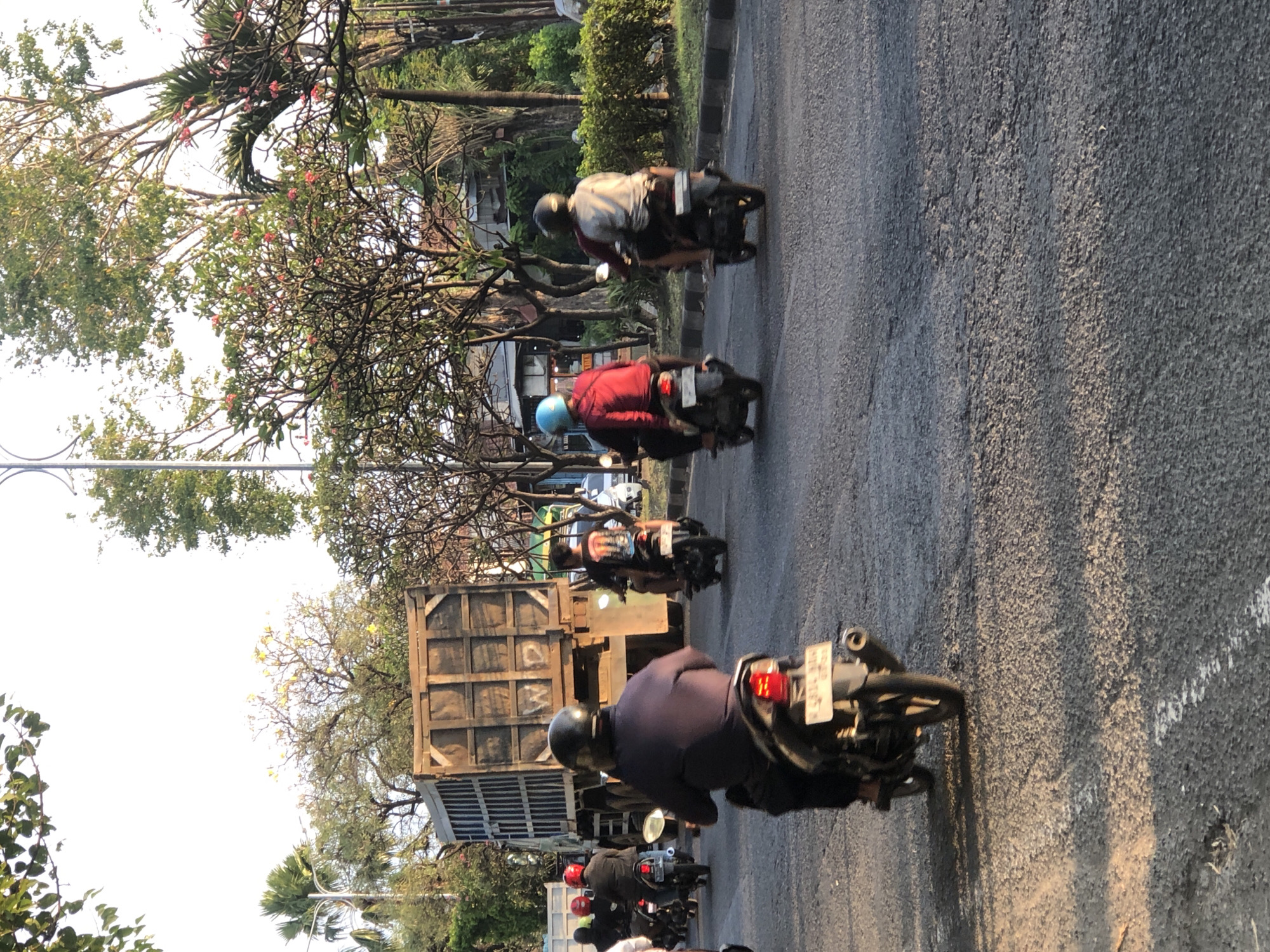 In Gelam, Indonesia, a truck with the letters "WP" painted on its rear sides is parked on the right side of a two-lane road. Several motorcyclists are riding on the road, with some in the foreground and others further back, approaching the truck. The scene is illuminated by sunlight, creating shadows on the road surface. The environment is lush with trees on both sides of the road, suggesting a warm climate. The sky is bright and clear, indicating daylight.