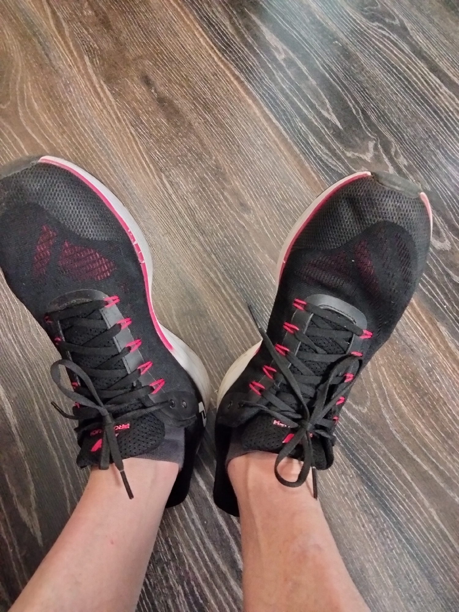 This is a close-up, top-down photograph of a person's feet wearing black athletic shoes with pink accents and laces, standing on a dark wood-grained floor. The shoes are tied loosely, and the socks are gray. The lighting is from above, casting subtle shadows. The environment does not provide any specific geographical indicators to determine its location within the city of Kyiv, Ukraine.