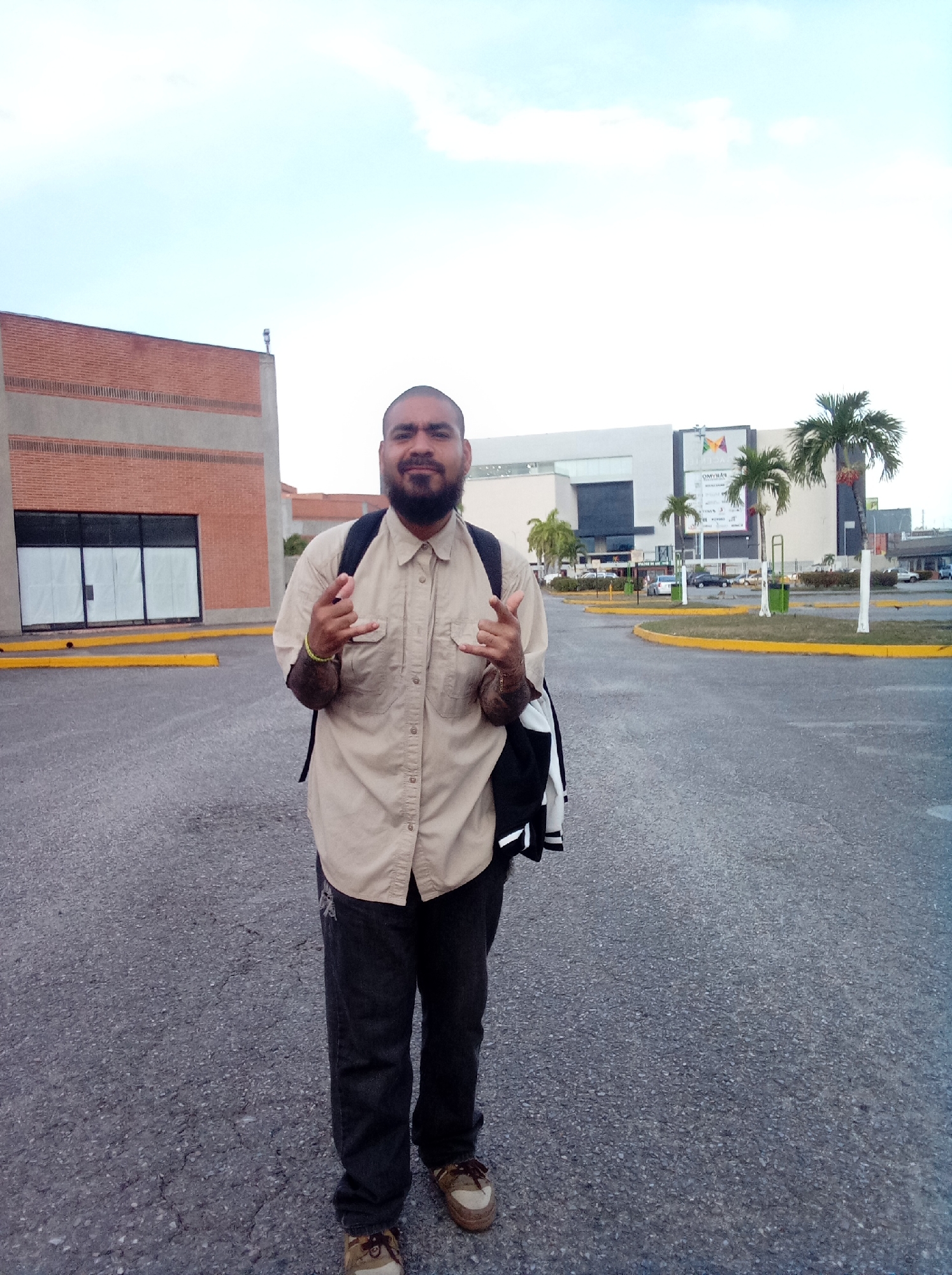 A medium shot depicts a man standing in a paved area in Parroquia Guatire, Venezuela. He is wearing a tan, button-down shirt, dark jeans, and sneakers. A backpack is slung over his shoulders. He is making a hand gesture with both hands. Behind him are buildings, including one with a brick facade, a large white building with a sign, and palm trees. The sky is overcast. The location appears to be a parking area or access road near a commercial building. There are painted curbs and landscaping features visible.