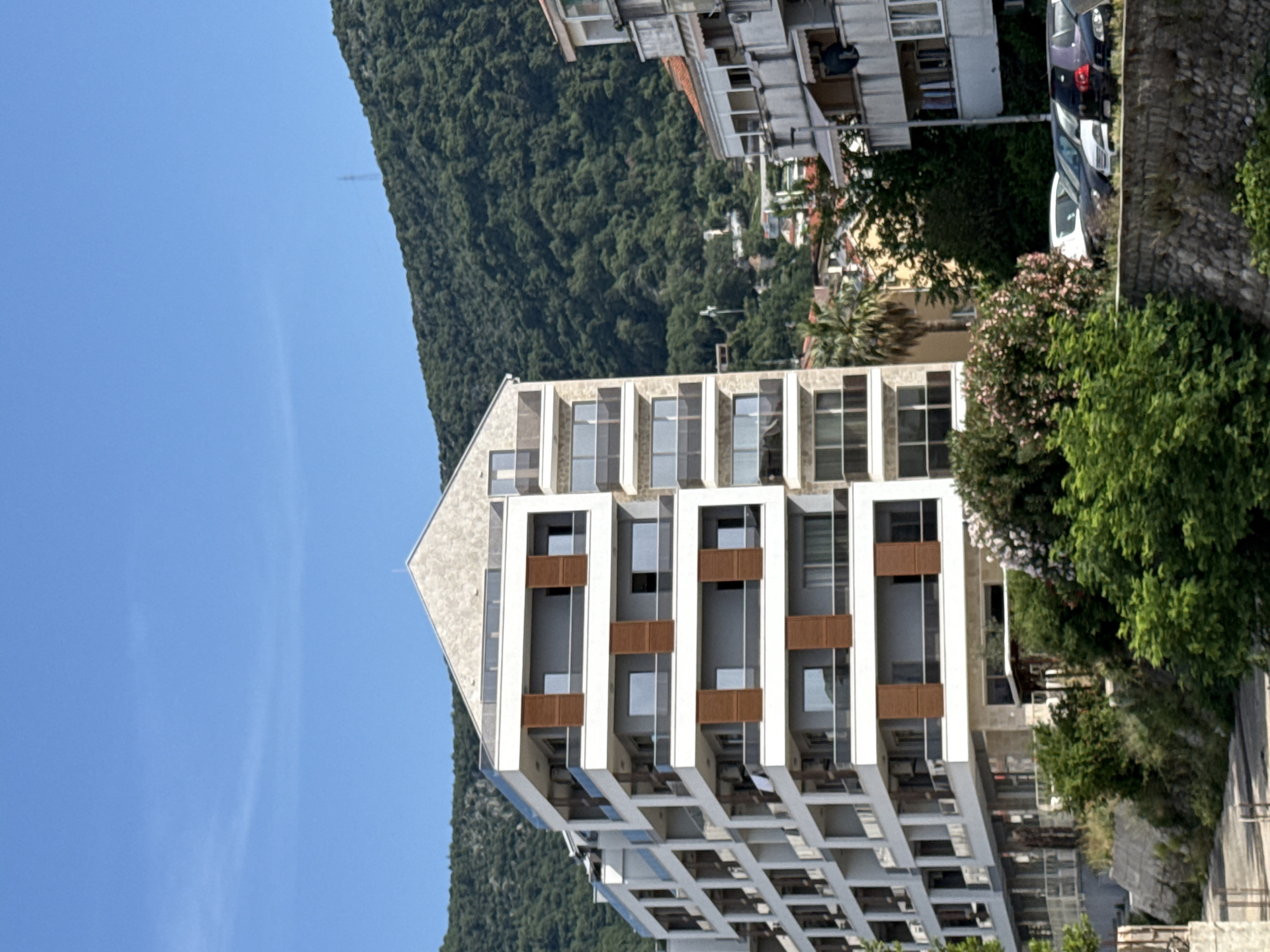 The image shows a view of a modern multi-story apartment building in Budva, Montenegro. The building is predominantly white with brown accents, and features large windows. The building is set against a backdrop of a lush green mountain, and a clear blue sky. Other buildings and some parked cars are visible in the background. A stone wall runs along the right side of the image. There is no sign of people in the shot. The image appears to be taken during daylight hours.