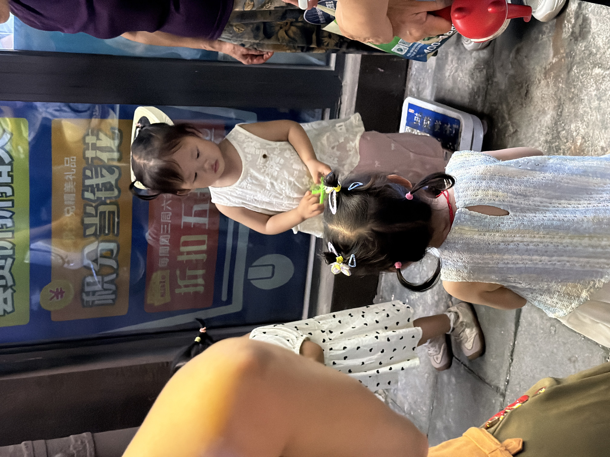 Observed in Chongqing High-tech Zone, China, is a scene with three young children. One child is wearing a white dress and is interacting with another child who is wearing a blue dress. The third child is wearing a black and white dress. The children appear to be on a city street next to a store. Adults are present in the periphery.
---