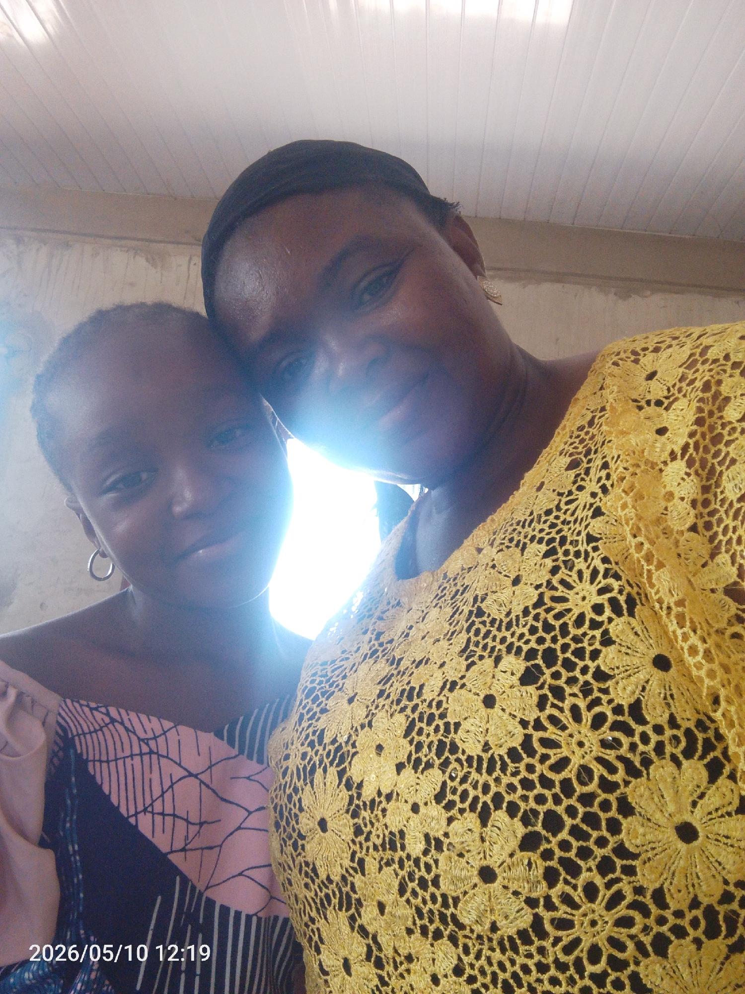 Two dark-skinned women pose closely together indoors. The woman on the right wears a yellow floral lace top. The woman on the left is wearing a patterned top with pink and black geometric shapes. The photo was taken on 2026/05/10 at 12:19. The location is Lokoja, Nigeria.
