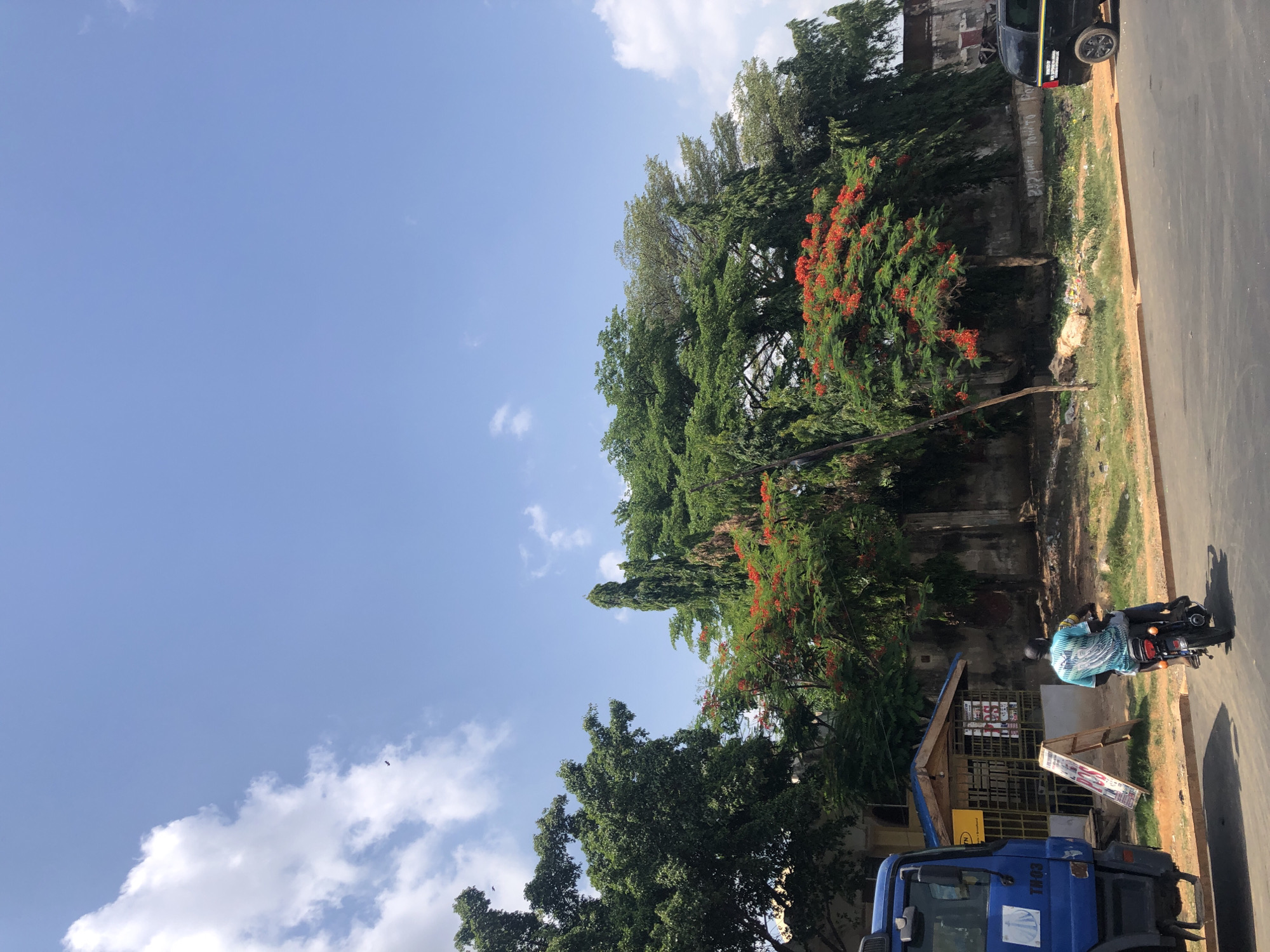 The image depicts a street scene in Akwanga, Nigeria. A blue truck is parked on the side of the road near a small shop. A person on a motorcycle is stopped next to the shop. Trees line the street, one with red flowers. A black vehicle is parked further up the road. The sky is blue with some clouds.