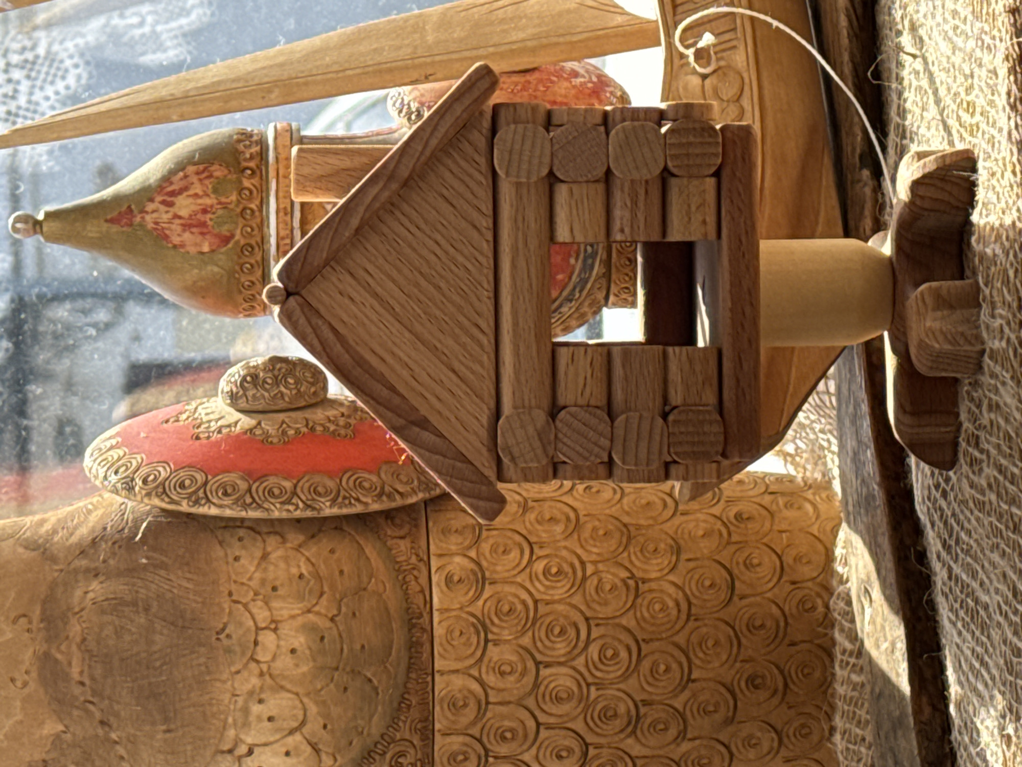 The image shows a collection of wooden objects. A wooden birdhouse with a pitched roof sits atop a turned wooden post, which is attached to a burlap-covered surface. To the left, a painted wooden object with a red lid and ornate carvings is visible. The background includes a window reflecting other objects, and a carved wooden surface with a spiral pattern. The location is Unknown, Unknown.
