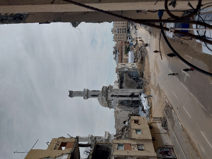 The image presents a wide-angle view of a street within a heavily damaged urban environment in Khan Yunis, Palestinian Territory, under an overcast sky. In the mid-ground, a large mosque with a tall, light-colored minaret stands prominently, displaying significant structural damage including partial collapses of its dome and main edifice, surrounded by extensive piles of rubble and debris. To the left of the mosque, a multi-story building is severely compromised, with large sections of its facade missing, exposing internal structures and rebar. A poster, possibly featuring cartoon characters, is visible on its lower level. The paved street, marked with white lines, extends into the distance, flanked by dirt shoulders and further debris. Approximately ten to fifteen individuals, primarily adults, are observed walking along the street and its shoulders, some singly and others in small groups, moving in various directions. In the immediate foreground on the right, a section of a damaged building with exposed rebar and a metal railing partially frames the scene. The background reveals additional multi-story buildings, some appearing less damaged than those closer to the foreground.