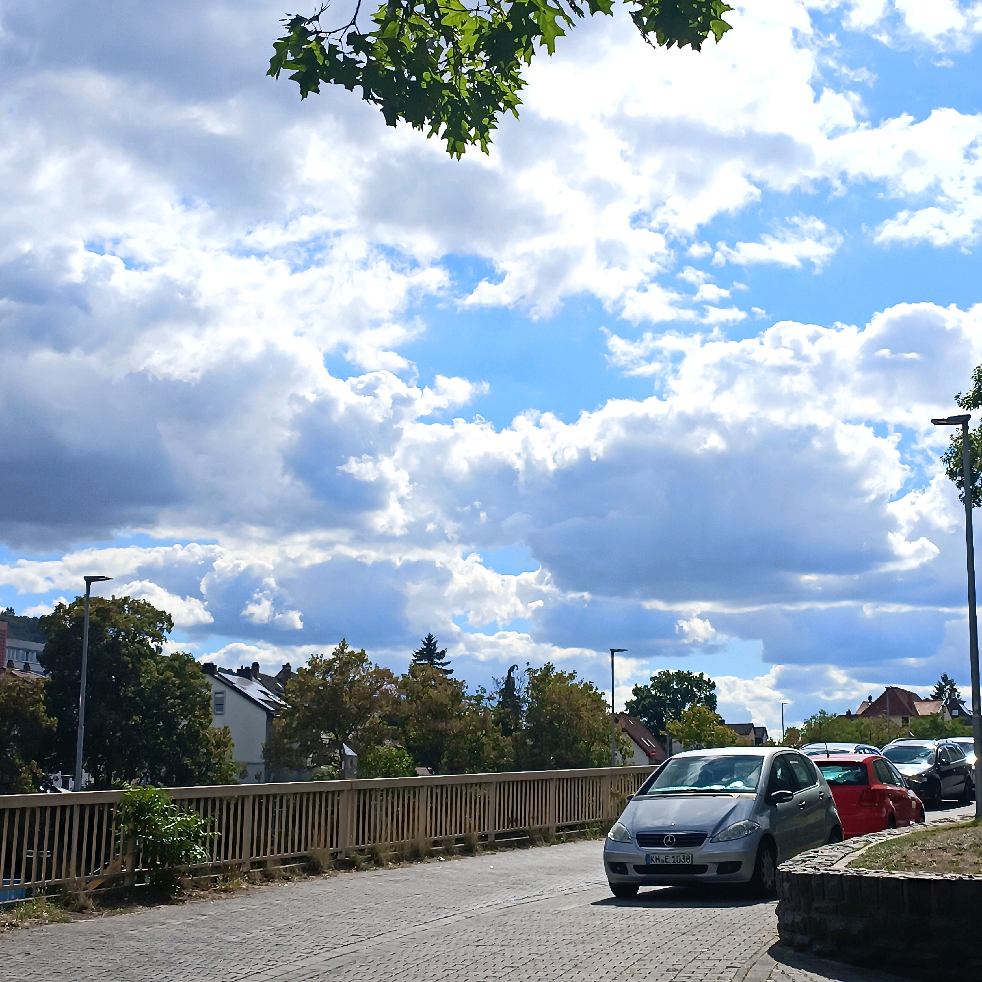 This is a street-level, eye-level, medium-angle, full-color photograph of a road in Bad Kreuznach, Germany, with cars and buildings visible. The sky is partly cloudy, with large, fluffy white clouds against a bright blue background. In the foreground, a gray Mercedes-Benz A-Class car is parked on the right side of the road. Its license plate reads "KH E 1038". To the left of the parked car, a red car is partially visible. Beyond the cars, a wooden fence with vertical slats runs along the side of the road. Behind the fence, trees and buildings with red-tiled roofs are visible. A streetlight is also present on the left side of the road. The road surface is paved with cobblestones. The overall impression is a typical urban scene on a pleasant day.