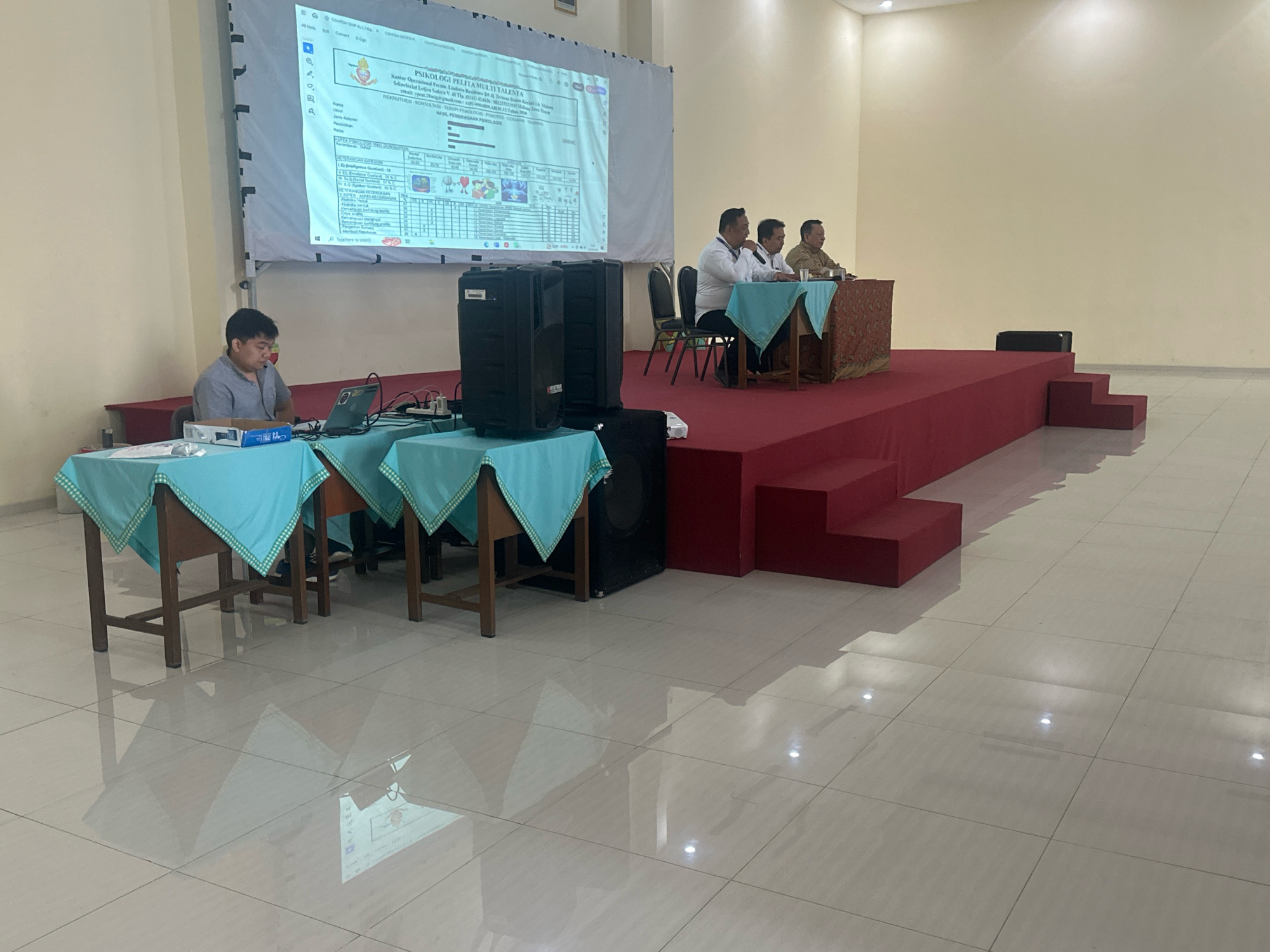 In a brightly lit, modern room with tiled flooring and cream-colored walls, a presentation is underway. A large projector screen displays a document with text and charts. Three men are seated on a raised, red-carpeted stage. Two men in white shirts are seated at a table draped with a blue tablecloth and a patterned runner, facing the screen. One of them is speaking into a microphone. A third man, in a brown shirt, sits at a separate table to the right, also draped with a blue tablecloth and a patterned runner. In the foreground, a young man wearing a grey polo shirt is seated at a table covered with a blue tablecloth, operating a laptop. Two large black speakers are positioned near his table. The room appears to be a conference or meeting space. There are no immediate visual cues to pinpoint the exact location within Mojokerto, Indonesia, beyond the context of a presentation being held in what seems to be a public or institutional building.