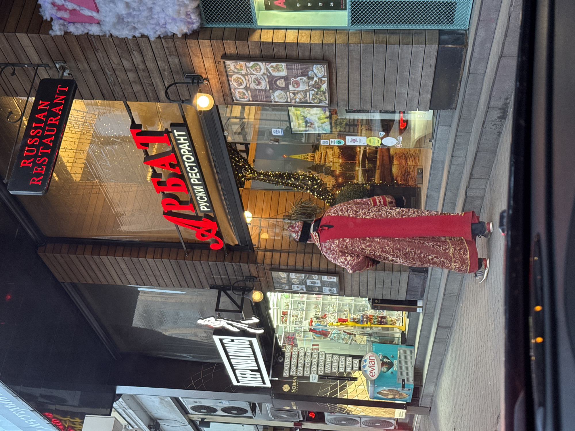 The image, rotated 90 degrees clockwise, depicts a lively street scene at dusk or night, likely in Sofia, Bulgaria. A person dressed in a striking, ornate costume walks past a restaurant and an adjacent shop.
The individual wears a long, patterned coat in shades of red and gold, possibly a traditional garment, along with a matching hat and modern sneakers. They are shown from behind, moving away from the restaurant's entrance.
The prominent establishment is a "Russian Restaurant," indicated by a black hanging sign with "RUSSIAN RESTAURANT" in illuminated red English letters. Below it, the restaurant's name, "АРБАТ," is displayed in large, illuminated red Cyrillic script, with "РУСКИ РЕСТОРАНТ" (Russian Restaurant) in smaller Cyrillic text underneath. The restaurant's facade features horizontal wooden paneling above large glass windows. Through the windows, string lights and a large mural depicting the Moscow Kremlin are visible, creating an atmospheric interior. A menu board showcasing various dishes is mounted on the wooden facade.
Adjacent to the restaurant, another shop front displays a neon sign reading "KEEP WALKING." Below this, a sign lists various items for sale: "SOUVENIRS," "SOBRA ESPRESSO," "CAPPUCCINO," "ICE CREAM," "CHOCOLATE," "COOKIES," "WATER," "CHIPS," "BEER," "NUTS," and "TEA," accompanied by an "evian" logo. Display cases inside this section hold various goods. The storefronts are brightly lit, indicating the evening or nighttime setting. The street features a paved walkway, and several small air conditioning units are visible at the bottom of the frame.