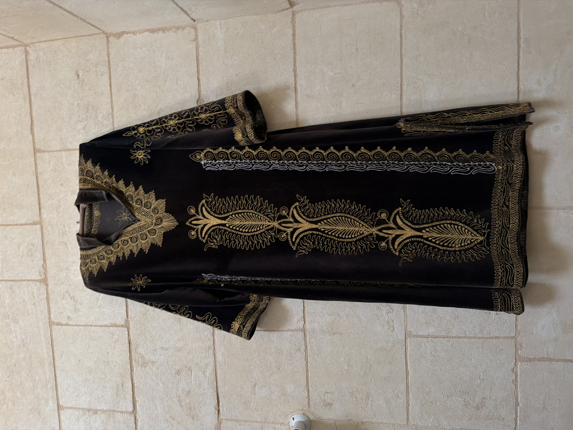 A dark brown velvet robe with intricate gold embroidery hangs on a wall constructed of light beige stone blocks. The robe has a V-neckline with embroidered detailing around the collar and down the front. The sleeves are short and also feature gold embroidery. Vertical embroidered panels with a paisley-like motif run down the front of the robe. The robe appears to be a traditional garment. The setting is an interior wall, possibly in Malta given the stone construction, though no specific location in Fontana, Malta can be identified from the image.