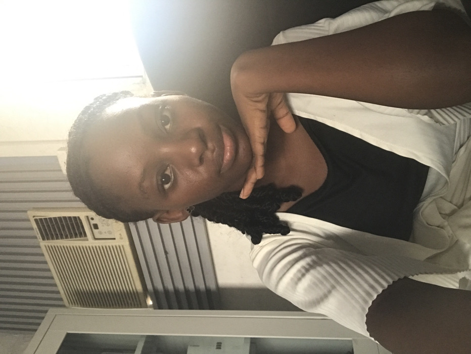 A chest-up portrait photograph depicts a young adult female positioned indoors in Ado, Nigeria. The individual, with dark skin and braided hair, looks directly forward. She wears a black top underneath a light-colored, pleated cardigan, and her right hand rests beneath her chin. In the background, to the left of the subject, a wall-mounted LG air conditioning unit is visible above horizontal window blinds. Partially visible further to the left is a light-colored cabinet with glass doors. The wall behind the subject features vertical striped wallpaper. Bright illumination emanates from the right side of the frame, highlighting the subject's right profile and the background area.