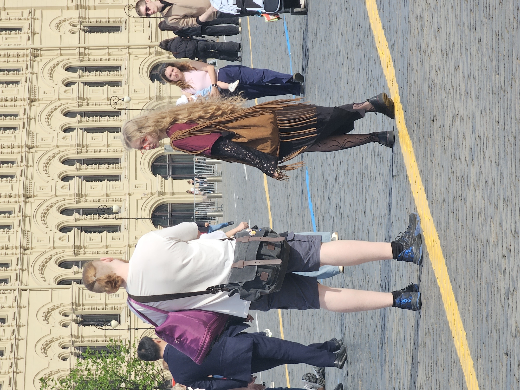 A daytime scene on a cobblestone square in Unknown, Unknown. A woman with long blonde hair, wearing a maroon shirt, fringed brown vest, black skirt, and patterned tights, stands near a yellow line on the pavement. Behind her is a light-colored building with arched windows. In the foreground, a person in a white shirt, gray shorts, and blue sneakers stands with a messenger bag over their shoulder. Other people are visible in the background, including a woman holding a baby. The environment includes cobblestone pavement, a building facade, and hints of greenery in the lower left.