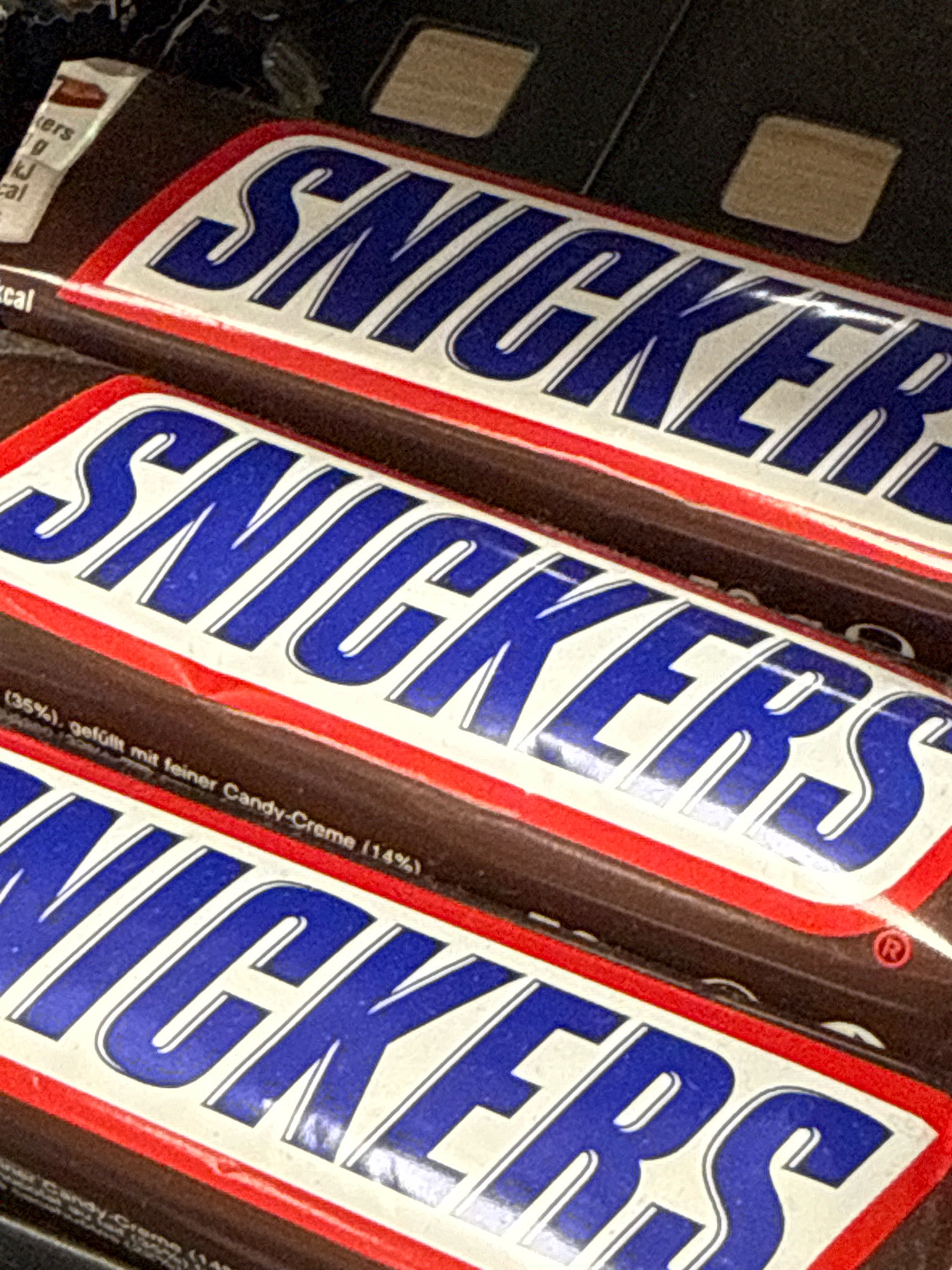 The image displays three Snickers candy bars arranged diagonally, with the top bar partially obscured by the one below it. Each bar features the "SNICKERS" logo in bold blue lettering on a white background, bordered by a red stripe. The bars appear to be stacked or displayed in a retail setting. Text in smaller font, likely ingredient or nutritional information, is visible on the lower part of the middle and bottom bars. The overall impression is a close-up shot of confectionery products. There is no indication of the city of Herzberg, Germany, or any specific environmental details that would suggest a particular location.