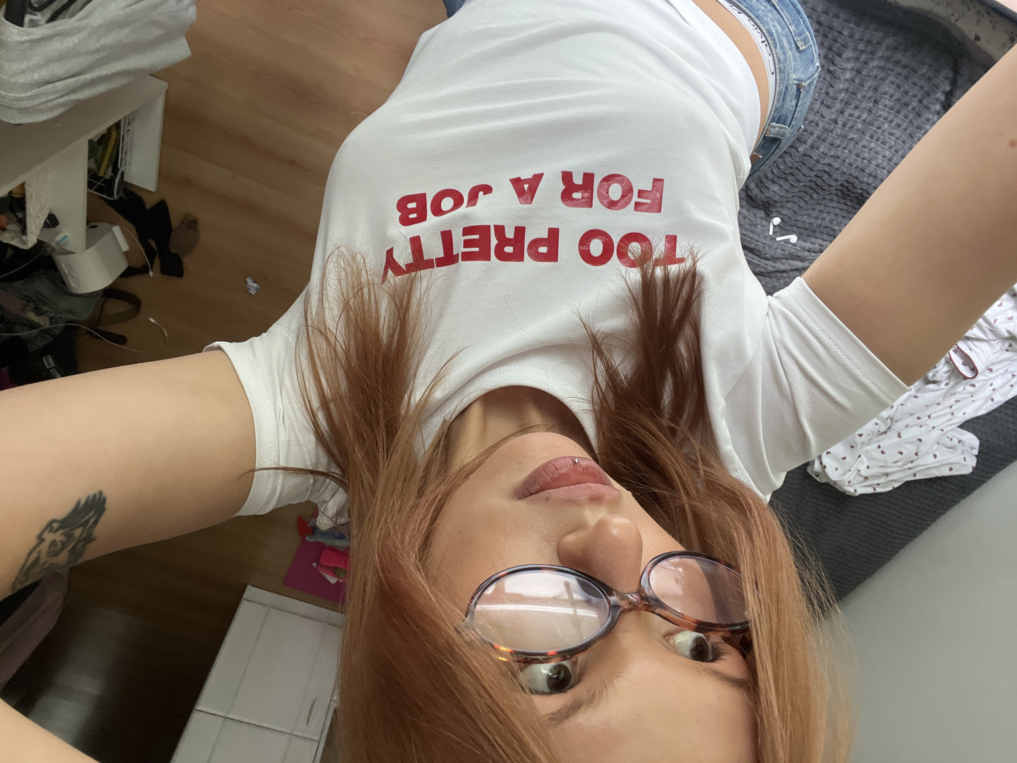 A medium-shot selfie captures a young person with long, reddish-brown hair and round, tortoiseshell-patterned glasses, lying down and looking upwards directly at the camera. They have a subtle lip piercing and a visible tattoo of a dark, winged figure on their left upper arm. The person is wearing a white t-shirt featuring the red, upside-down text "TOO PRETTY FOR A JOB" across the chest. Blue denim shorts or jeans are visible, with the white waistband of "Calvin Klein" underwear peeking out above them on their left hip.
The setting appears to be an indoor room in Chaikovsky, Russia. The background shows a light wooden floor to the left, cluttered with various items including a white low table or shelf, cables, and dark fabric. To the right, a dark grey textured blanket or bedspread covers a surface, with white wireless earphones (AirPods) resting on it. A folded white garment with small red heart patterns is also visible on the blanket. The overall scene conveys a casual and relaxed atmosphere.