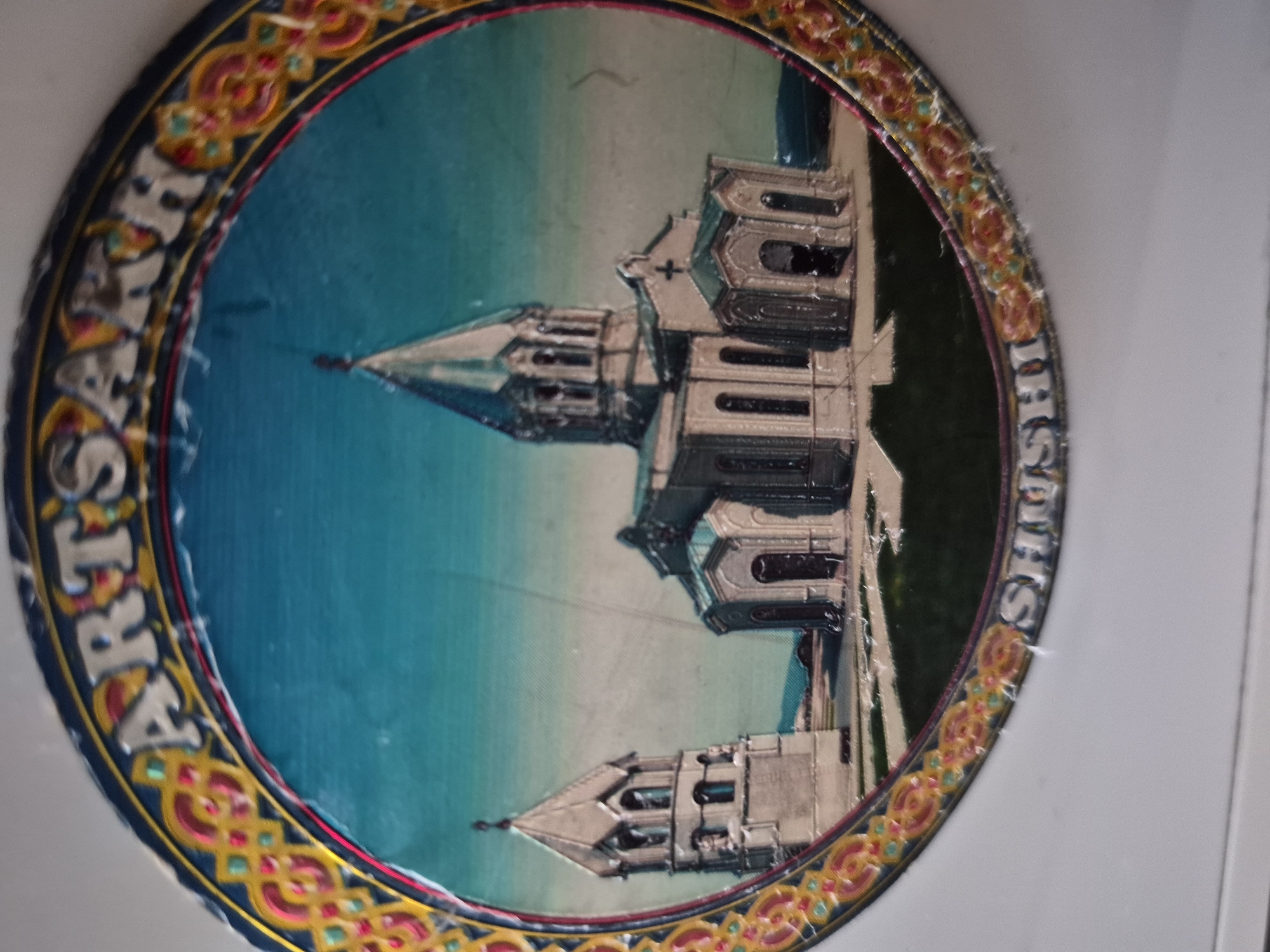 The image is a close-up, slightly angled shot of a decorative round plate or plaque featuring a depiction of a church with a bell tower, set against a teal sky. The artwork is enclosed within a decorative border with Armenian-inspired motifs in red, yellow, and blue. The words "ARTSAKH" are visible in silver lettering along the top arc of the border, and "SHUSHI" is visible in similar lettering along the bottom arc. The church appears to be a stone structure with a central dome and a separate bell tower. The overall scene is rendered in a somewhat stylized, almost illustrative manner. The plate itself shows signs of wear, with some minor scuffing and peeling of the decorative elements. The background of the image is a plain, light-colored surface, likely a wall or table. It is important to note that this is a depiction on a plate, not a direct photograph of the actual location. The location of the depicted church, if it is indeed a real landmark, is not directly verifiable from the image itself, and the claim that it is in Vanadzor, Armenia, is not supported by the visual information provided.