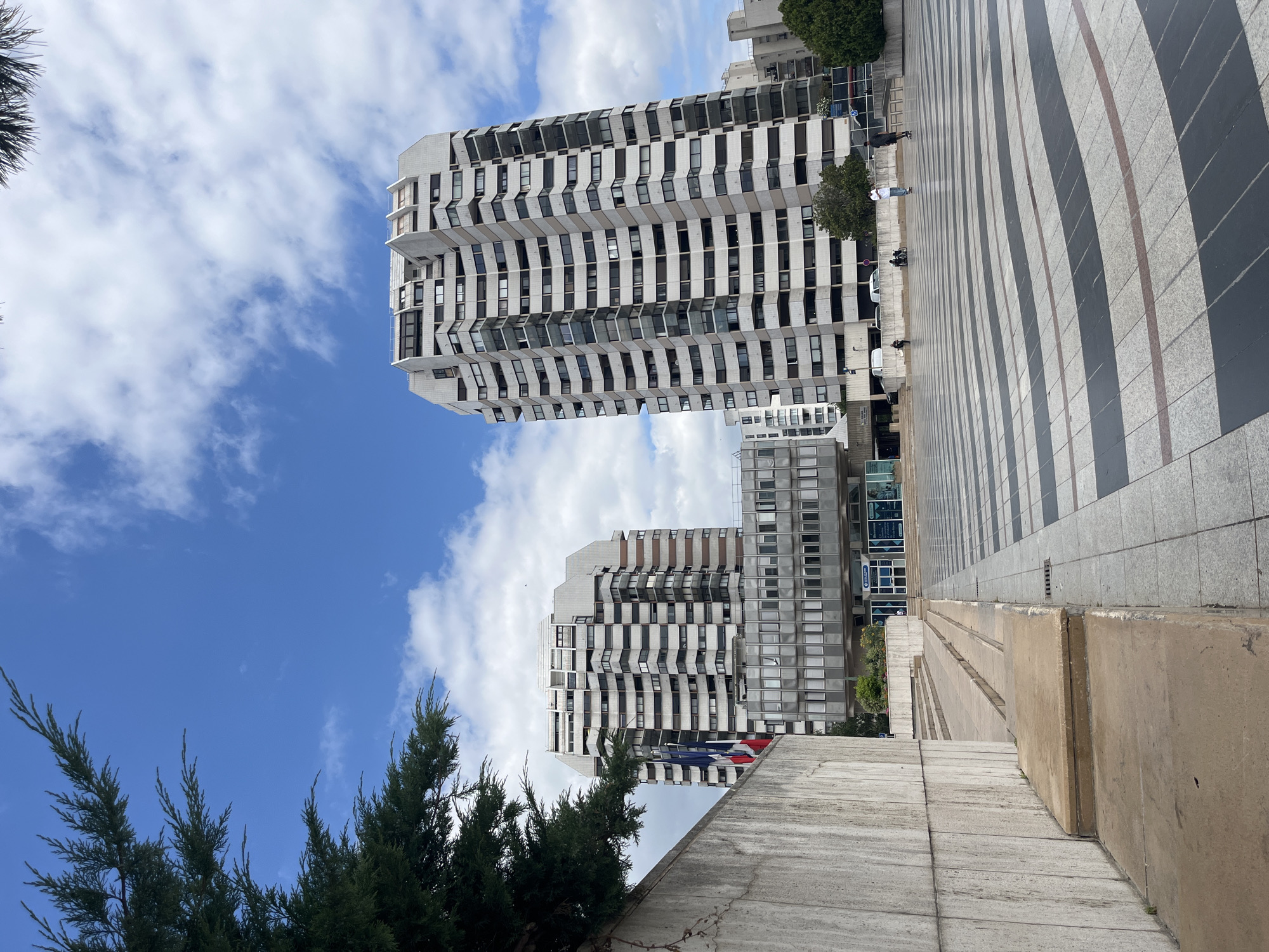 This is a wide shot of a plaza in Créteil, France, dominated by two tall, modern apartment buildings with white, staggered facades. The plaza itself features a striking black and white striped paving pattern. In the foreground to the left is a large, rough-textured concrete wall. To the right of the wall, concrete steps ascend. In the mid-ground, a lower, rectangular building with a facade of reflective glass windows and horizontal white mullions sits between the two taller buildings. The sky above is a bright blue with scattered white clouds. A few people are visible in the plaza, one person is skateboarding. A tree with dark green foliage is visible in the upper left corner. The French flag is also visible, attached to a pole on the left side of the frame.