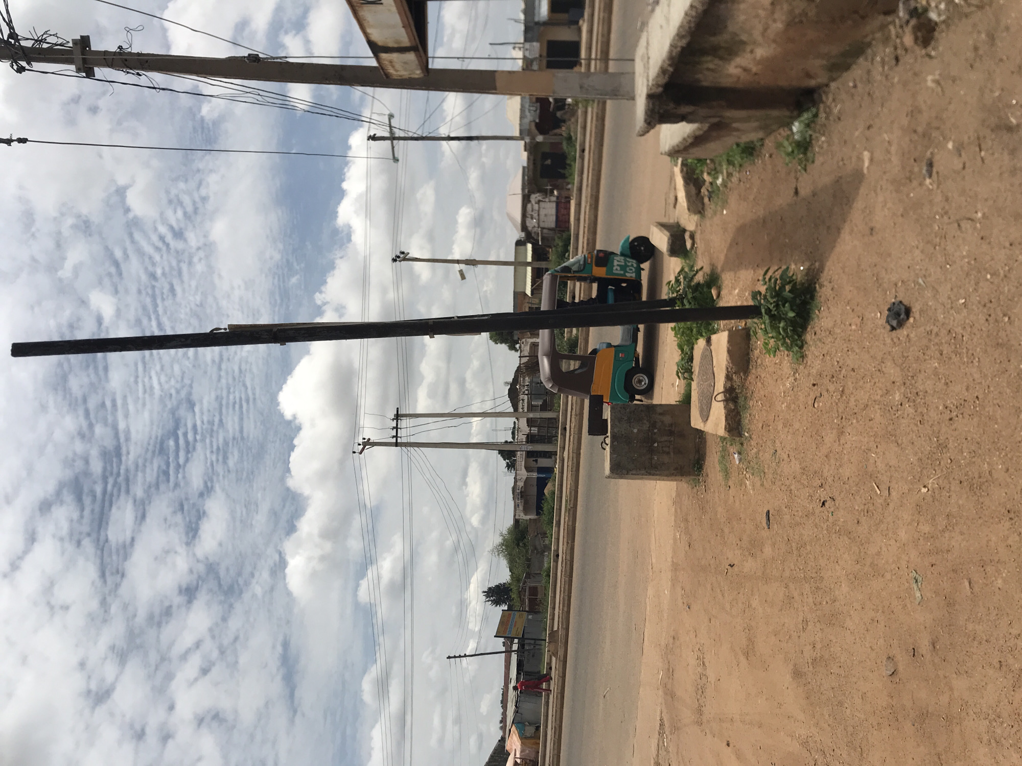 This image captures a street scene in Rafin Par, Nigeria, under a bright, partly cloudy sky. The foreground features reddish-brown laterite soil, which forms a dirt path alongside the main asphalt road. Scattered debris, including plastic and small pieces of trash, is visible on the ground. A tall, black utility pole stands prominently in the center-right foreground, casting a long shadow towards the left. To its right, a concrete low wall or barrier runs parallel to the path.
In the midground, an asphalt road curves slightly. A small, three-wheeled vehicle, commonly known as a "keke napep" or tuk-tuk, is parked or moving slowly on the right side of the road. It is distinctively colored in light blue/green and yellow, with the alphanumeric "PTA 10 S" visible on its rear. On the opposite side of the road, a person dressed in a bright red outfit walks leftward on a dirt verge.
Numerous utility poles, some wooden and others concrete, crisscross the scene, supporting a complex network of overhead electrical and communication wires against the sky. Behind the walking person and the left side of the road, there are various informal structures, including market stalls with corrugated iron roofs and tarpaulins, alongside more permanent but modest buildings. Further down the road on the right, more single-story buildings line the street, some appearing weathered or under construction. The background shows a continuation of this semi-urban landscape with distant trees and structures under the expansive cloudy sky.