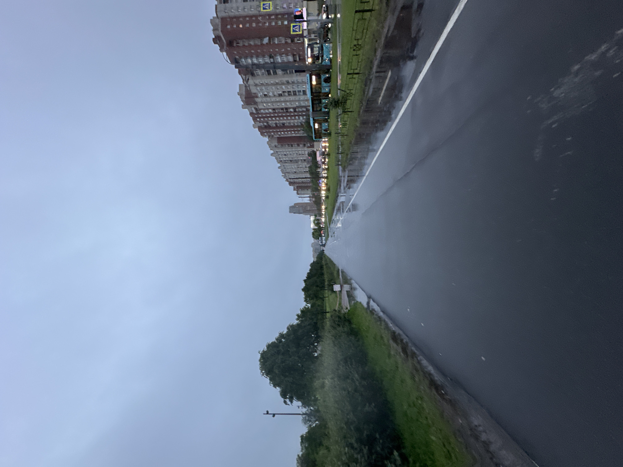 The image depicts a wet, overcast day in Saint Petersburg, Russia. A wet asphalt road, reflecting the cloudy sky, stretches into the distance. On the left side of the road, there is a grassy area with trees and bushes. On the right side, a row of multi-story residential buildings with brick and concrete facades line the street. There are several buses on the road, and streetlights are visible in the distance. The presence of puddles on the road and the overcast sky indicate recent or ongoing rain. The general atmosphere is muted and somber due to the weather conditions.