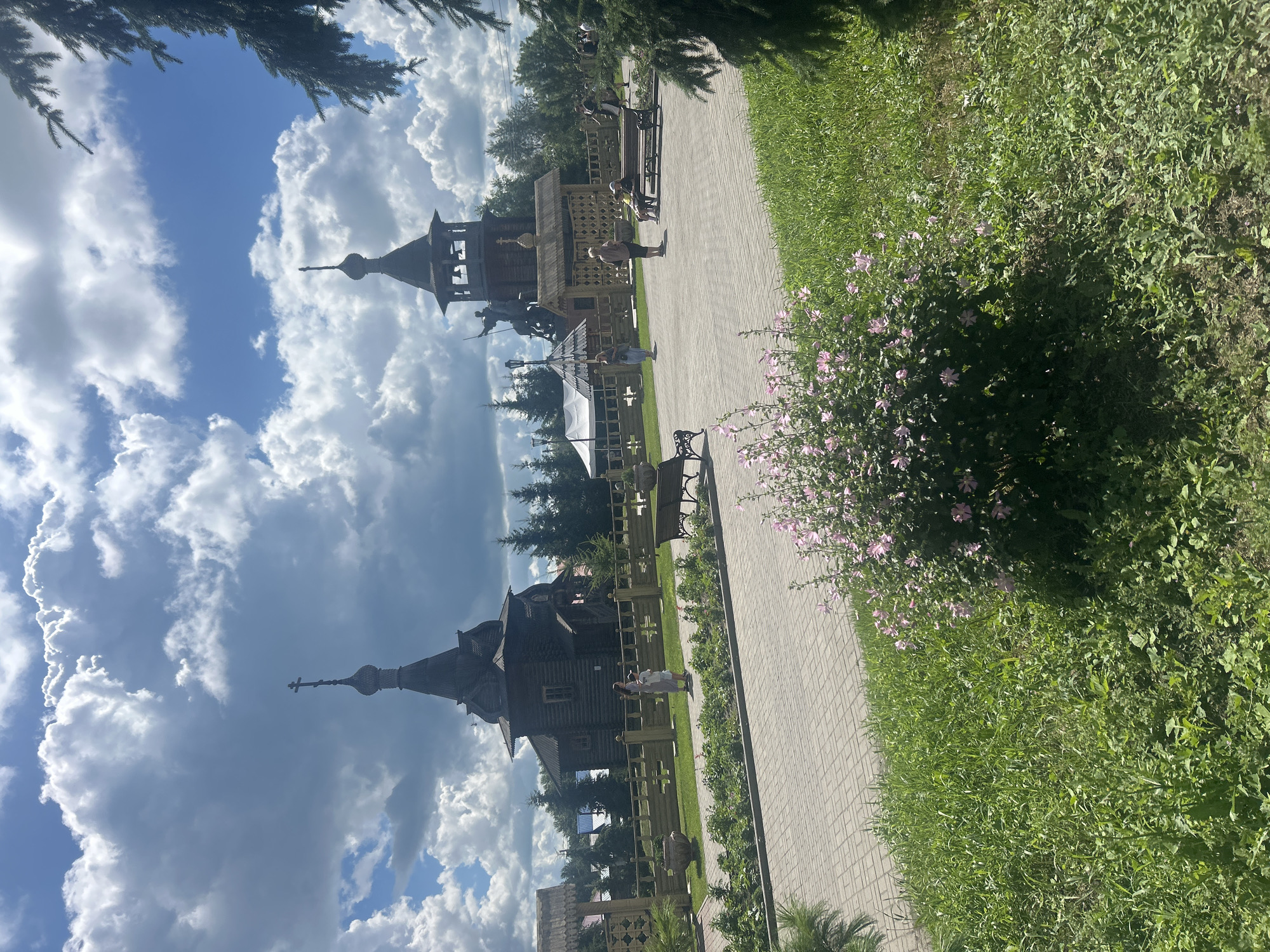 The image depicts an outdoor scene in the city of Bolsherechye, Russia, featuring a complex of wooden structures including two churches, a bell tower with a statue of a horseman, and a gatehouse. The architecture is distinctly traditional Russian. In the foreground, a paved walkway runs through a grassy area with a bush of pink flowers. People are visible in the middle ground, some walking, others sitting on benches. The sky above is partly cloudy with large, fluffy white clouds against a bright blue backdrop. The overall impression is of a cultural or historical site during daytime.
