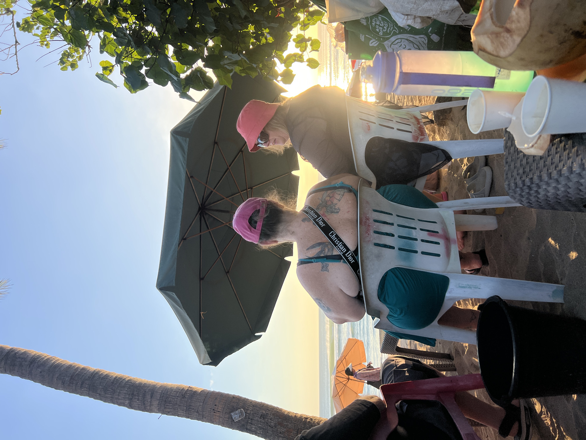 Two women are seated on plastic chairs on a sandy beach in Kuta, Indonesia, facing the ocean at sunset. The woman on the left wears a pink baseball cap and a teal bra with tattoos on her upper back and shoulder. The woman on the right wears a pink bucket hat and sunglasses, her gaze directed towards the setting sun. They are partially shaded by a large green umbrella. In the foreground, a black bucket and various cups and a coconut are visible on a woven table. In the background, a palm tree stands to the left, and lush green foliage to the right. Another umbrella shades a person walking on the beach further down. The sun casts a warm glow across the water.