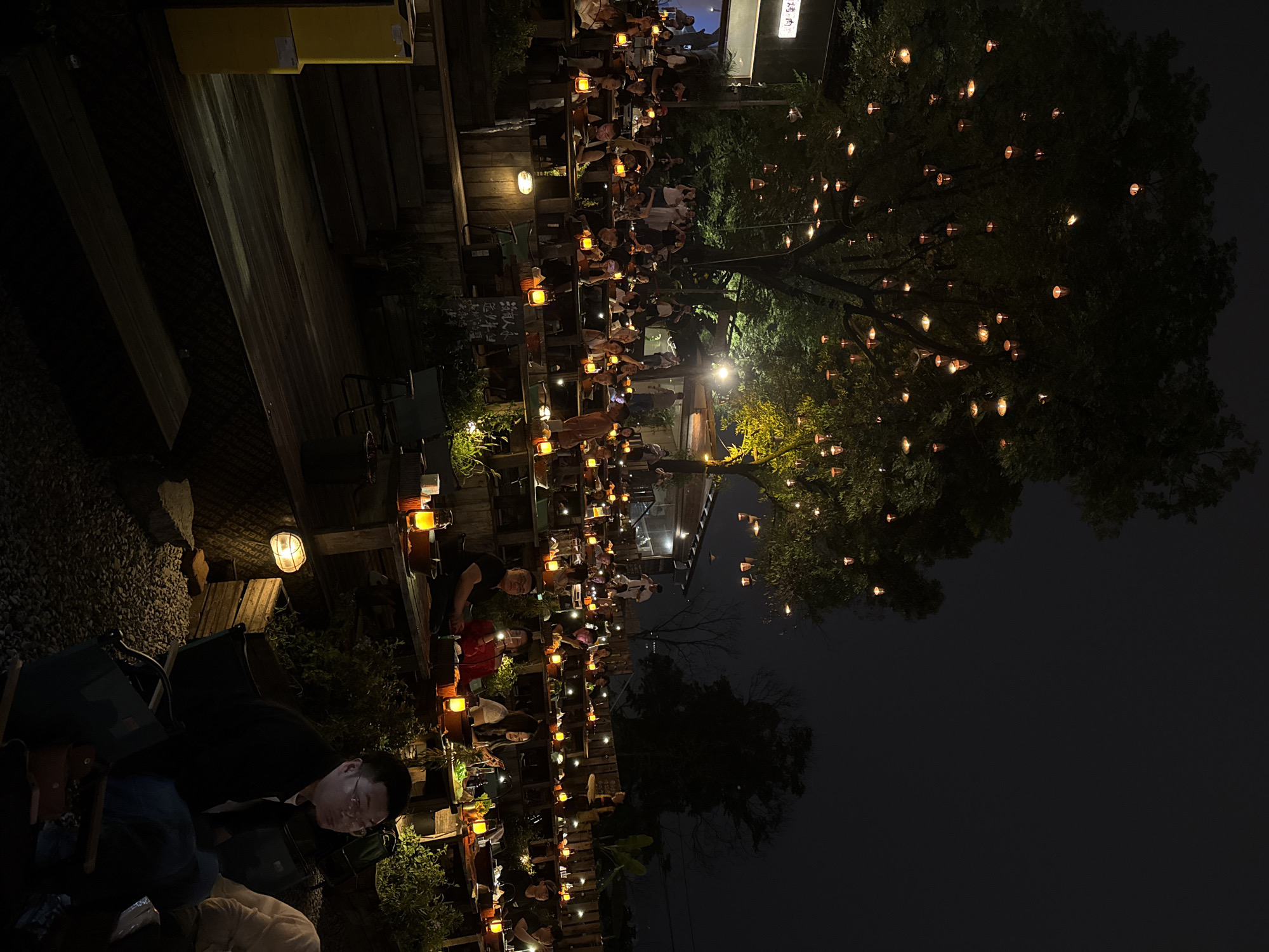 The image shows an outdoor dining area at night, likely in the Nan'an District, China. Multiple rows of wooden tables are occupied by people, illuminated by small lanterns placed along each table. A large tree in the background is also decorated with lanterns. In the foreground, a person is seated, and other furniture, possibly chairs or bags, are visible. The area is dimly lit, and the overall atmosphere appears festive.