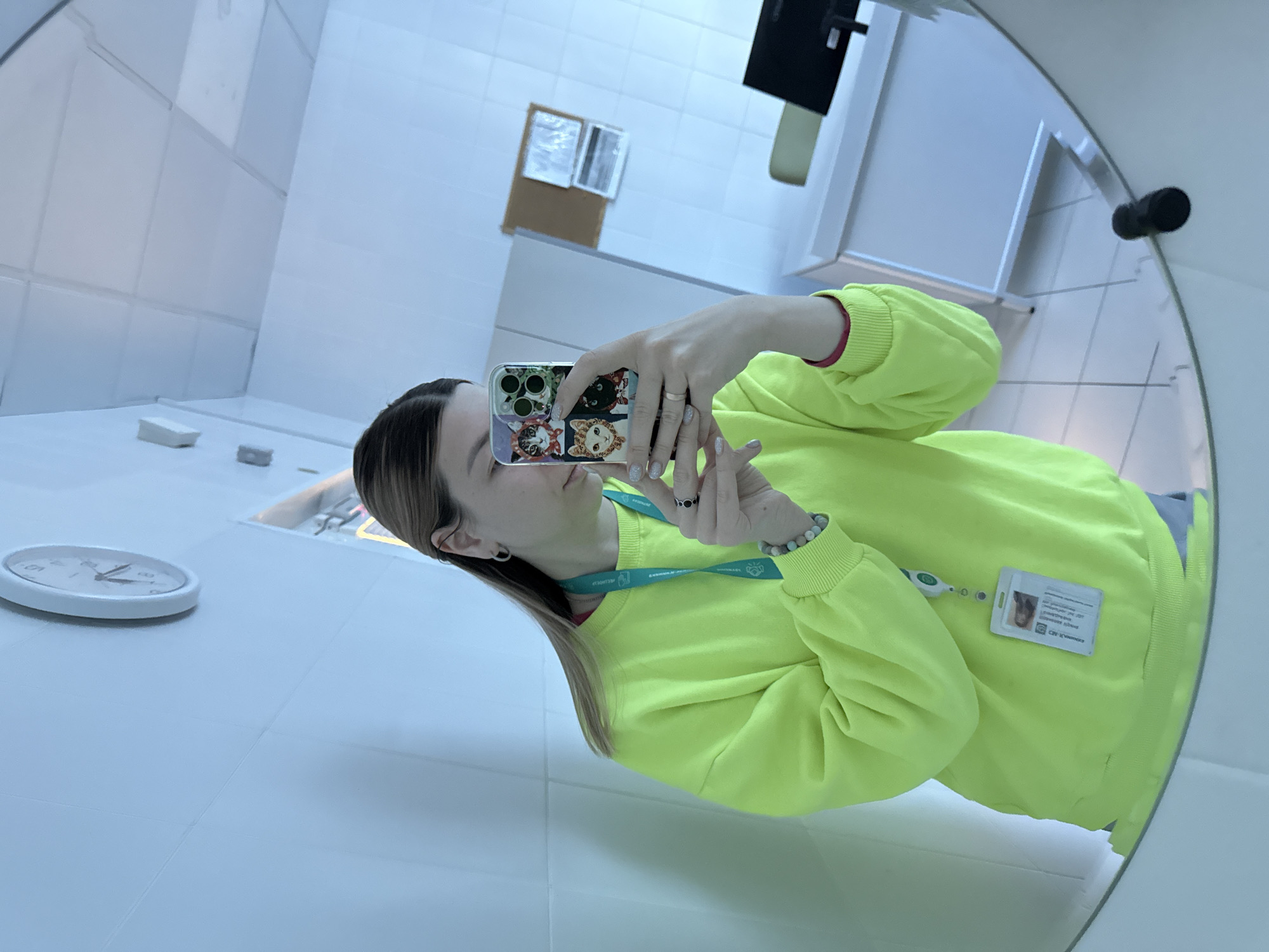 A woman in a bright yellow sweater is taking a selfie in a room with white tile walls. She is holding a phone with a cat-themed case. She is wearing a lanyard with an ID badge. A clock is visible in the lower left corner. The scene appears to be in the "Zaryadye" park media center in Moscow, Russia, due to the distinctive interior design.