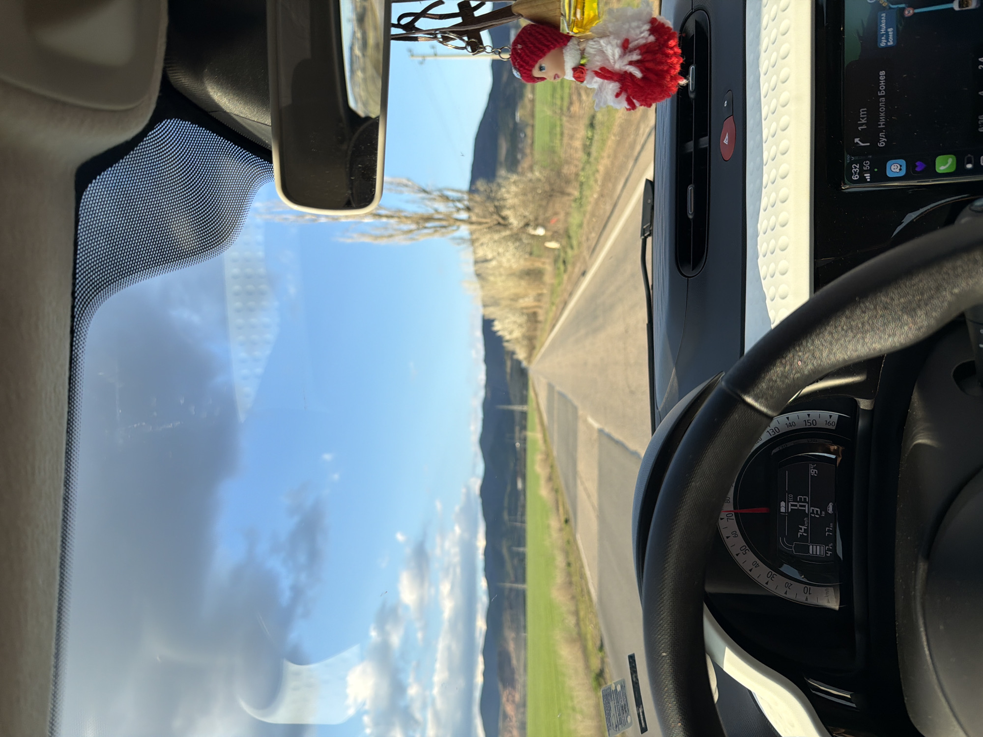 The image shows a driver's perspective from inside a vehicle, traveling on a paved road in Buhovo, Bulgaria. The speedometer indicates a speed of 74 km/h. A mobile phone mounted on the dashboard displays a navigation app showing directions to "бул. Никола Бонев" which is approximately 1 km away, and the time is 6:32 AM. The weather outside appears sunny with scattered clouds and a clear blue sky. The landscape features fields and distant hills. A small decorative doll hangs from the rearview mirror.