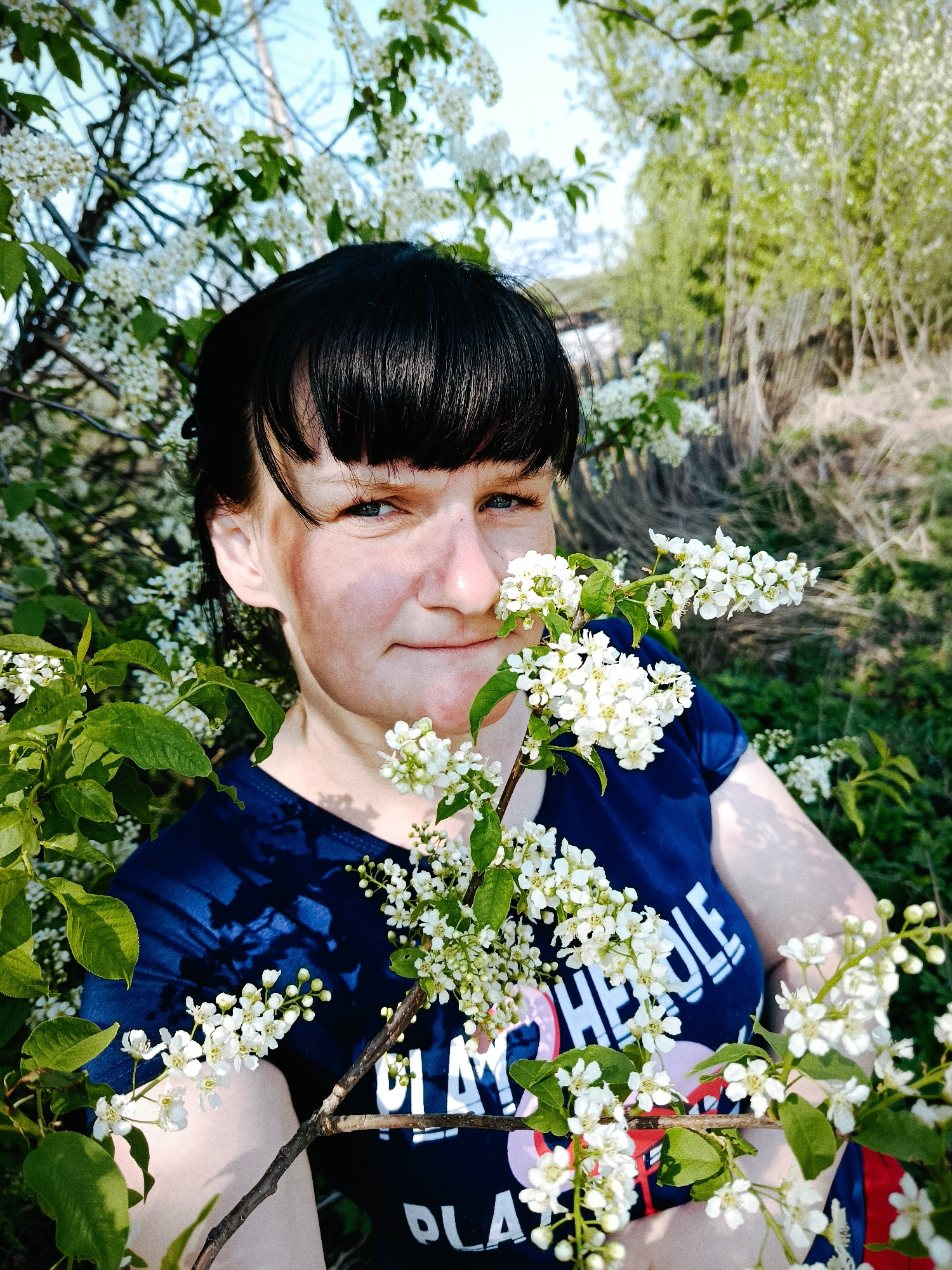 A woman with dark hair and bangs is standing among flowering branches, likely in a natural setting within the city of Фоки, Russia. She is wearing a navy blue t-shirt with white lettering. The background consists of trees and vegetation.