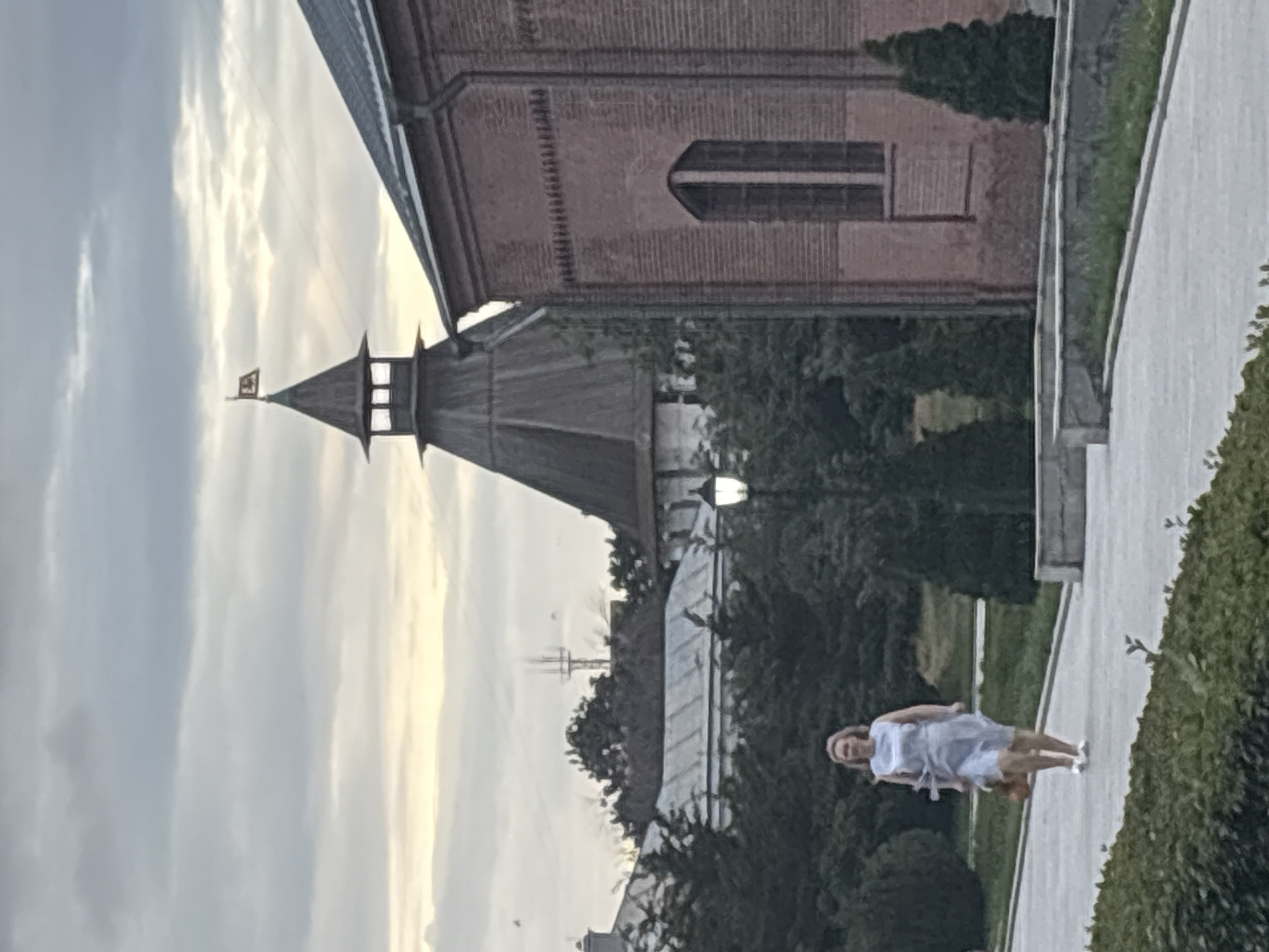 The image depicts an outdoor scene within the Astrakhan Kremlin complex in Astrakhan, Russia, under an overcast sky. In the mid-ground, slightly right of center, stands the multi-tiered Cathedral Bell Tower (Prechistenskaya Gate Tower) with a dark wooden conical roof culminating in a spire topped by a small flag displaying a dark emblem on a light field. To the immediate right of the tower, a red brick building with a dark roof and a prominent downspout features a tall, narrow arched window with dark framing. The brickwork includes a decorative corbeling pattern near the roofline.
In the foreground, a light-colored paved pathway extends diagonally from the lower left towards the mid-right. A blonde woman of apparent middle age is walking on this path from left to right, facing slightly towards the camera. She is wearing a light blue sleeveless dress and white or light-colored flat shoes. She carries a light brown, possibly woven, bag in her right hand.
Between the walking woman and the brick building, a black lamppost with a white illuminated lantern is visible. Lush dark green trees and manicured bushes frame the pathway and partially obscure the lower sections of the buildings. In the far background, behind the main tower, other buildings with light-colored roofs are visible, along with a tall, slender antenna or cell tower structure. The sky is predominantly gray with scattered white clouds; a faint band of lighter, yellowish sky is present on the horizon beneath the clouds. A few dark specks, possibly birds, are visible in the upper sky above the background foliage.