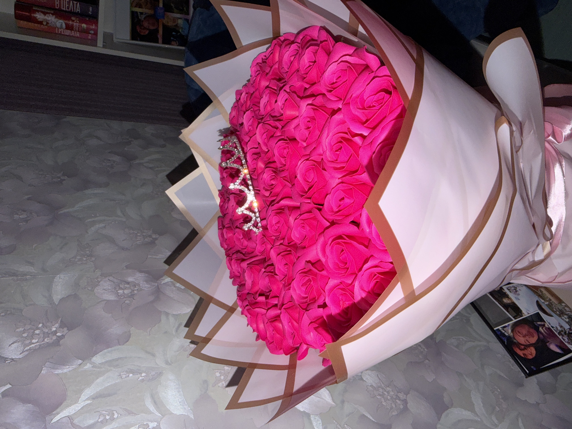 A bouquet of fuchsia-colored roses wrapped in white and gold paper with a small, jeweled tiara placed on the roses, sits on a surface with a floral pattern. There are books in the background. Based on available information, the image location is unknown and the image does not have enough context to determine if the location is in Burgas, Bulgaria.