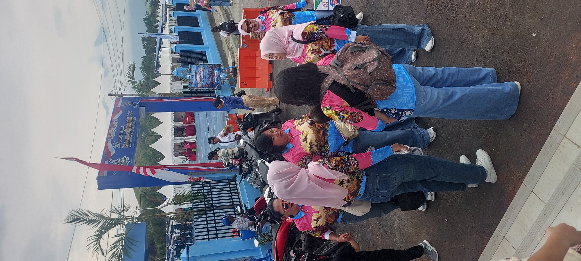 The image depicts an outdoor gathering in front of a blue arched entranceway, likely at a festival or event, in Asei Besar, Indonesia. Several people, primarily women wearing matching brightly colored shirts with a tropical bird and landscape design and blue jeans, are standing in groups. Some are wearing hijabs. One woman is holding a baby. Motorcycles are parked nearby. In the background, white tents are set up, suggesting vendor stalls or event facilities. A banner above the entrance reads "FESTIVAL DANAU SANTANI XV". The weather appears overcast.