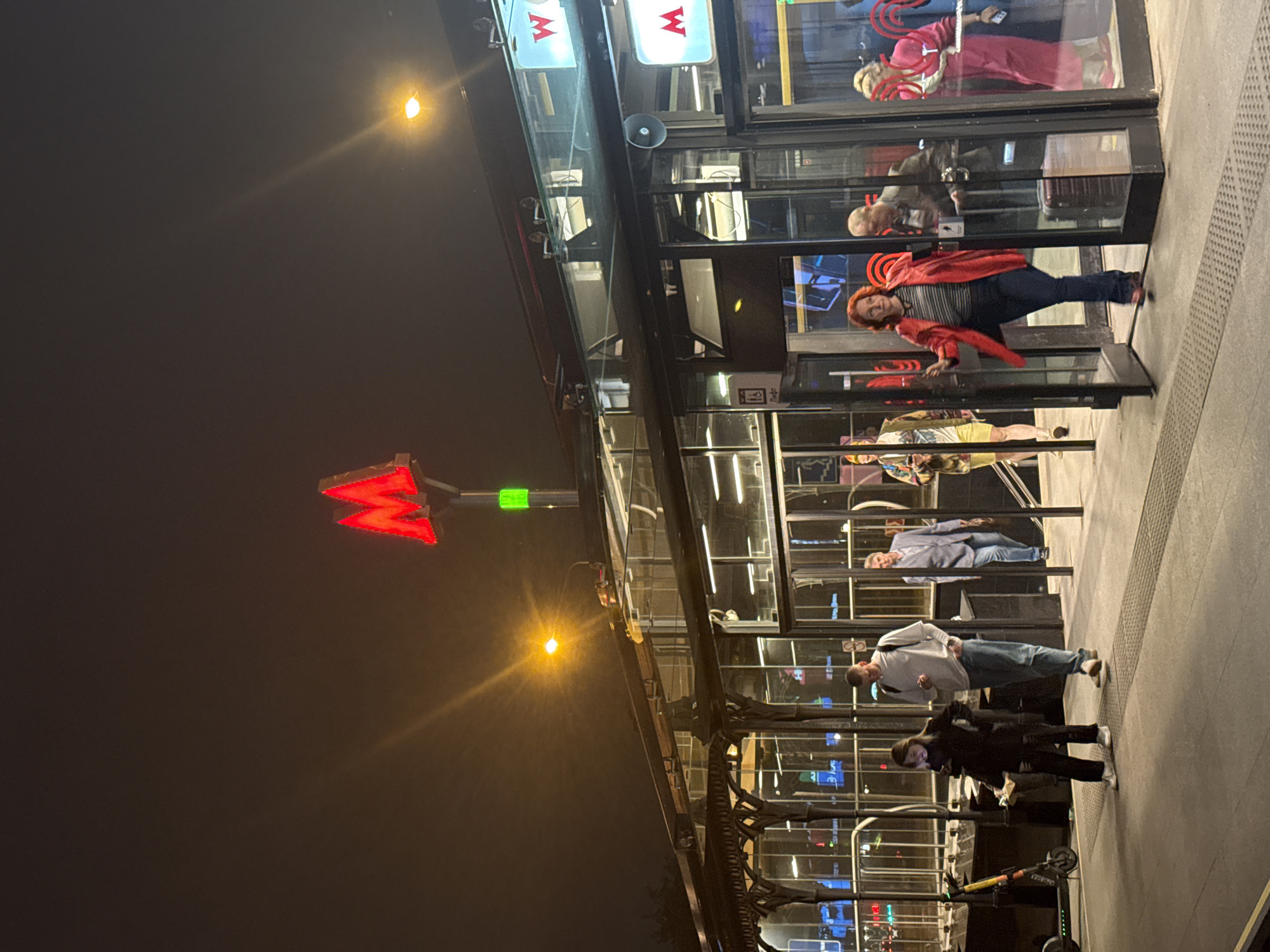 The image depicts a Moscow Metro station entrance at night. A red "M" logo is prominently displayed. Several people are visible inside and near the entrance, with a person in a red jacket about to enter. Scooters are parked nearby. Street lights and the glow from the station illuminate the surrounding area.