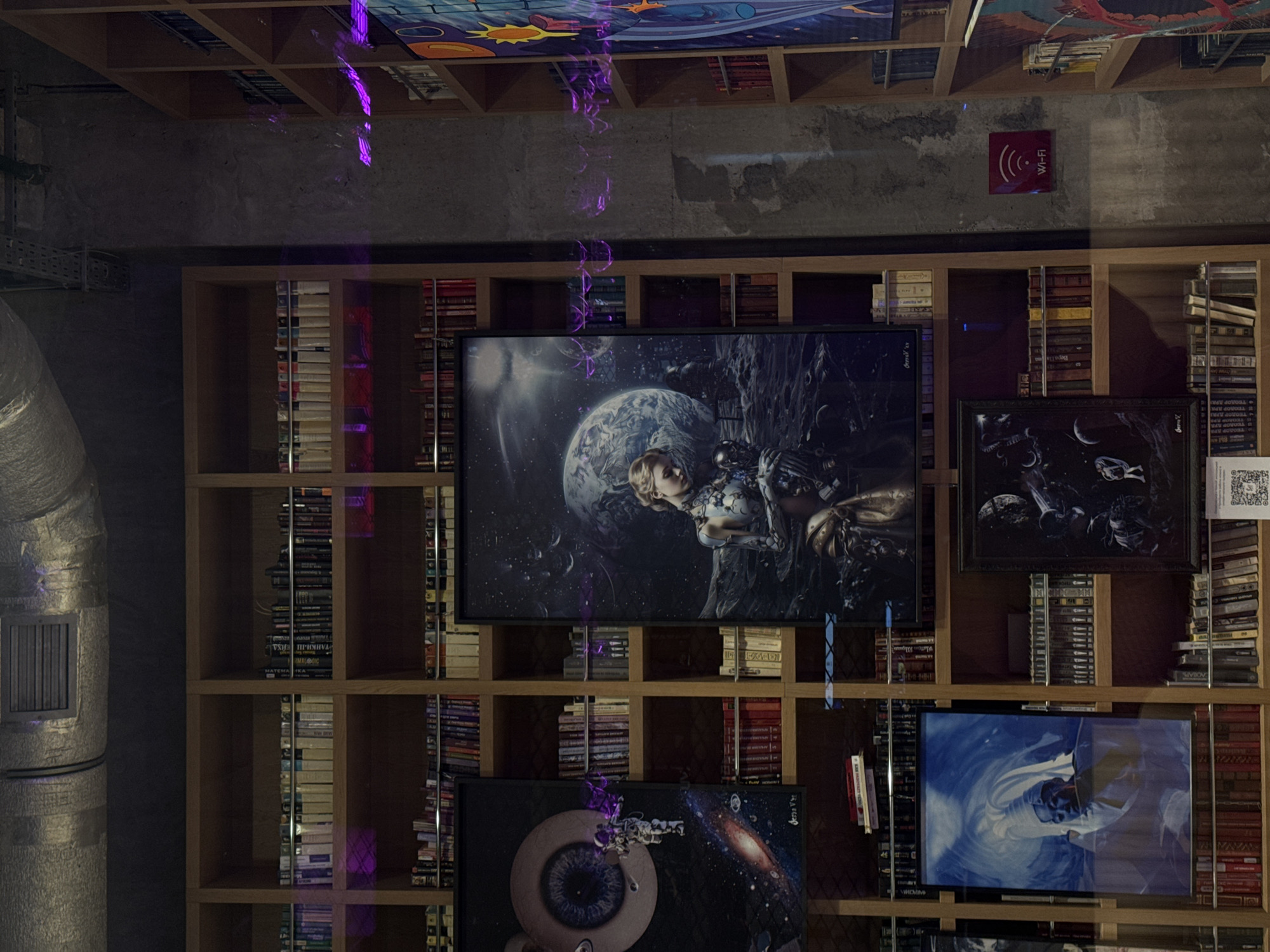 The image shows an interior space with a wooden shelving unit filled with books and various framed artworks. The shelving unit appears to be against a wall with a textured, possibly concrete, finish. A Wi-Fi sign is visible on the upper right side of the wall. A large cylindrical duct runs vertically along the left side of the image.
The artwork includes images related to space, science fiction, and fantasy themes. One artwork prominently features an astronaut or figure in futuristic attire with a celestial body in the background. Another shows a galaxy-like image.
The lighting is somewhat dim and artificial, with purple light streaks across the image, likely from a light source.
Without further details, it's challenging to pinpoint the precise location in Moscow. Additional information about the establishment would be required to determine its location accurately.