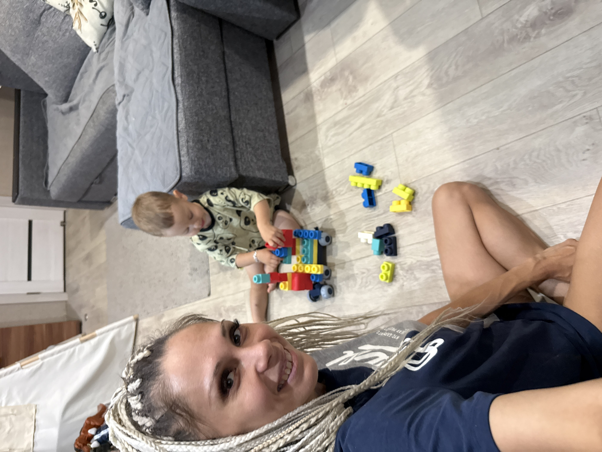 In a domestic interior, likely a living room, a woman with blonde braided hair sits on a light-colored wooden floor, smiling at the camera. She is wearing a dark blue t-shirt. Beside her, a young child, appearing to be a toddler, is engaged in playing with colorful building blocks, constructing a train-like structure. Scattered on the floor are additional building blocks, some yellow and blue. In the background, a grey sectional sofa is visible, along with a child's teepee structure and a doorway. The lighting suggests an indoor setting during the daytime. The specific location of Rusanovo, Russia, is not visually ascertainable from the image.