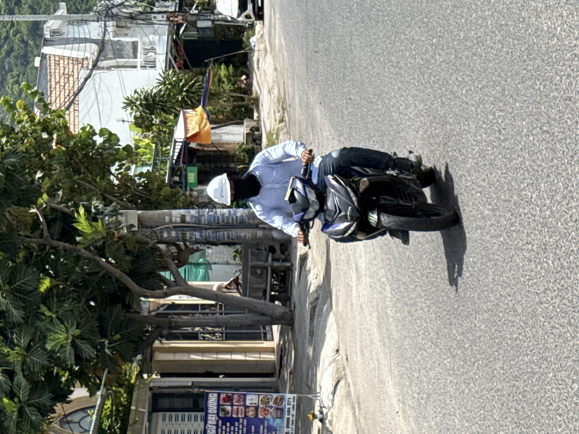 A man wearing a white hard hat and a black face covering rides a black and blue motorcycle down a paved road. He is wearing a light blue long-sleeved shirt and dark pants. The motorcycle has a blue and white design on its front. The man is positioned in the center of the frame, riding towards the viewer.
The road is an asphalt surface with visible texture. To the left of the road, there is a sidewalk area with some trees and buildings. A sign with Vietnamese text and images of food is visible on the left side of the sidewalk. Behind the man, more buildings and trees line the street, with a hillside in the background. The sky is clear and the scene is brightly lit, suggesting daytime. The location is identified as Bac Nha Trang Ward, Vietnam.