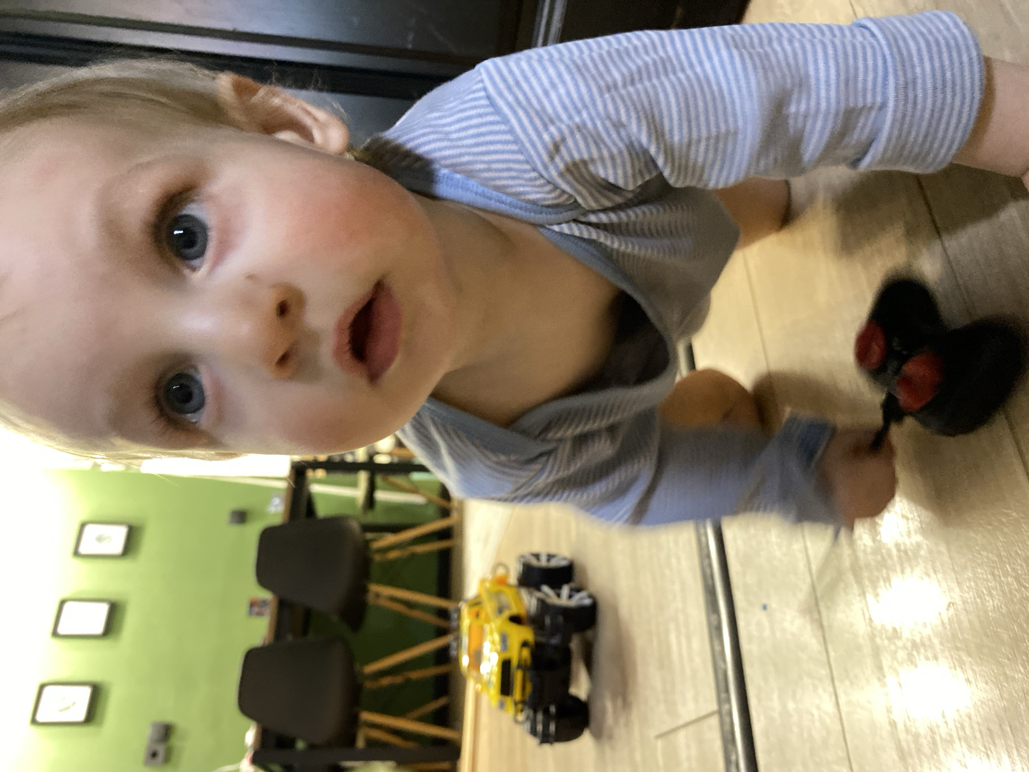 A child with light hair and blue eyes, wearing a light blue and white striped shirt, is on a light-colored floor. The child is looking up at the camera with their mouth slightly open. A black and red toy is in the child's right hand. Behind the child is a yellow and black toy truck and two black chairs. A green wall with three framed pictures and an electrical outlet is visible in the background. The location cannot be determined from the image.