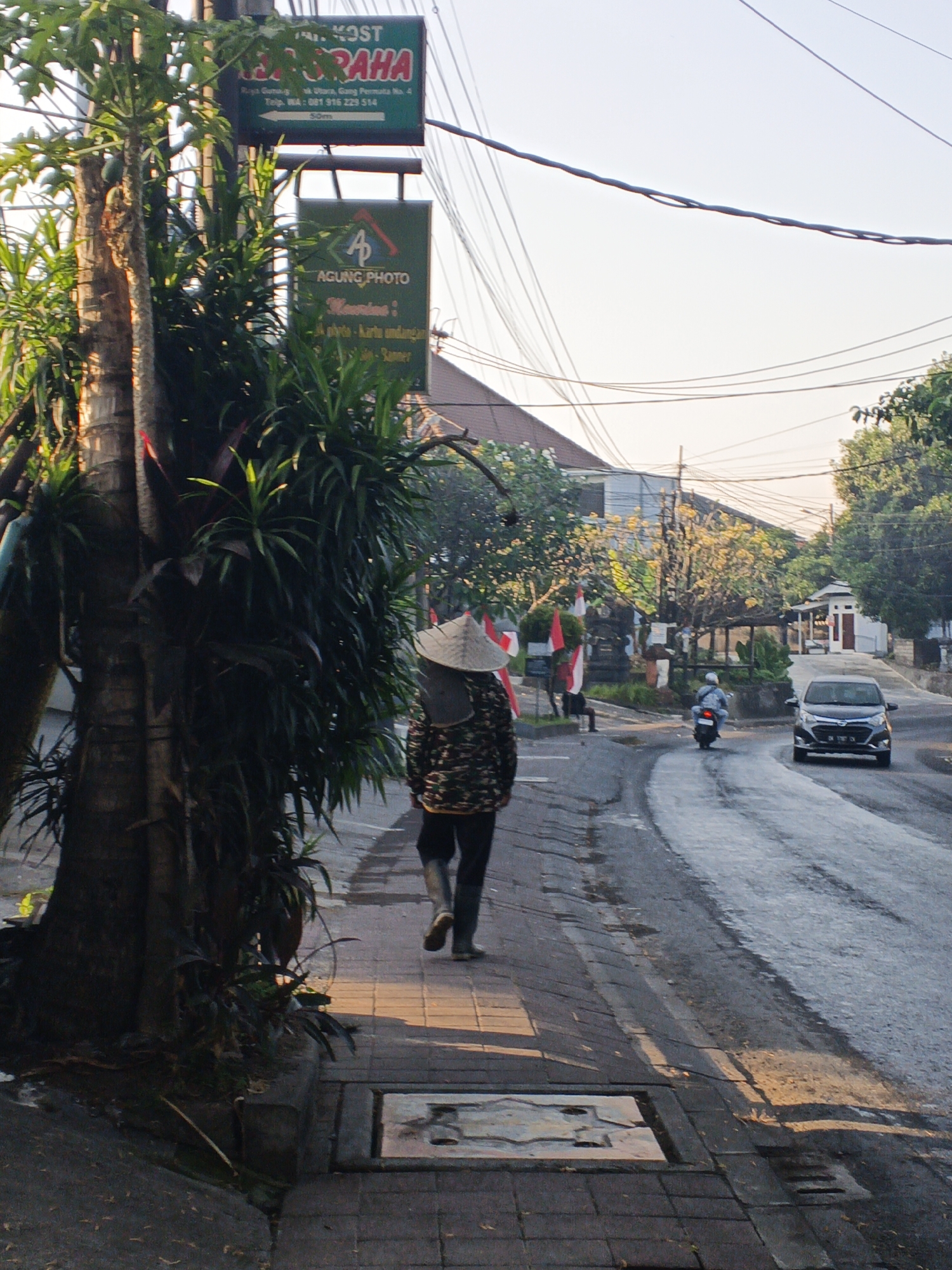 The image depicts a street scene in North Kuta, Indonesia, at what appears to be late afternoon or early morning due to the long shadows. A person wearing a conical hat and a patterned jacket is walking on a paved sidewalk. The sidewalk is bordered by a road where a black car is driving away from the camera and a motorbike is approaching. On the left side of the image, there is a large tree with dense foliage. There are also street signs visible, including one for "KOST JRAHA" and another for "AGUNG PHOTO." Buildings and lush greenery are present in the background, with power lines crisscrossing the sky.