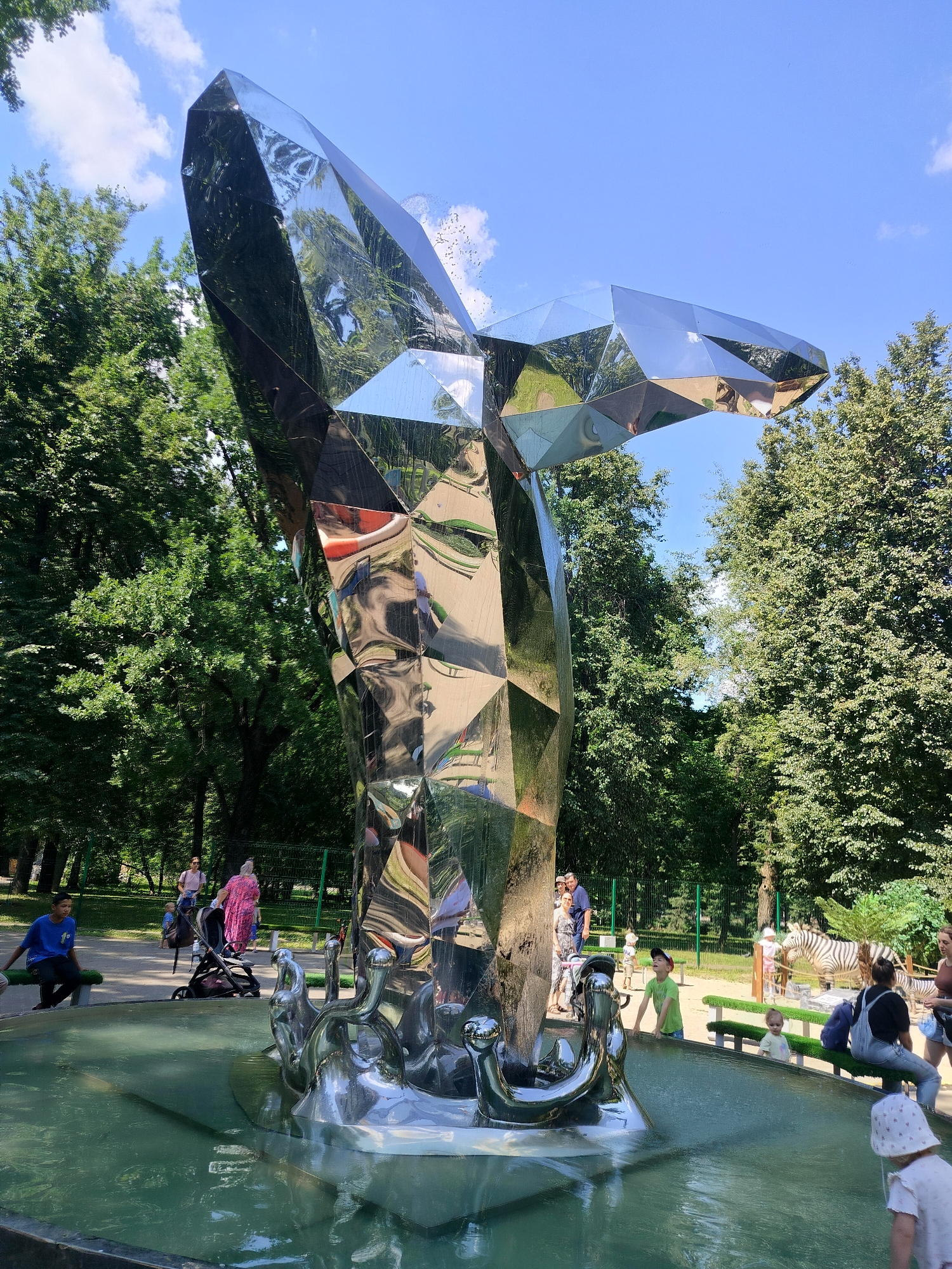 The image shows a large, abstract, geometric sculpture made of polished metal, resembling a crystal or a stylized whale tail, situated in a park with lush green trees under a clear blue sky. The sculpture is part of a fountain, with water spraying around its base, creating reflections on its polished surface. Several people are visible in the park, including adults and children. In the background, there are benches, a pathway, and a green fence. To the right of the sculpture, a zebra exhibit is visible in the distance, suggesting the park might be a zoo or a part of a zoo. The overall setting indicates a sunny day in a public outdoor space. The sculpture appears to be located in a park in Moscow, Russia, likely in a public area or a zoo.