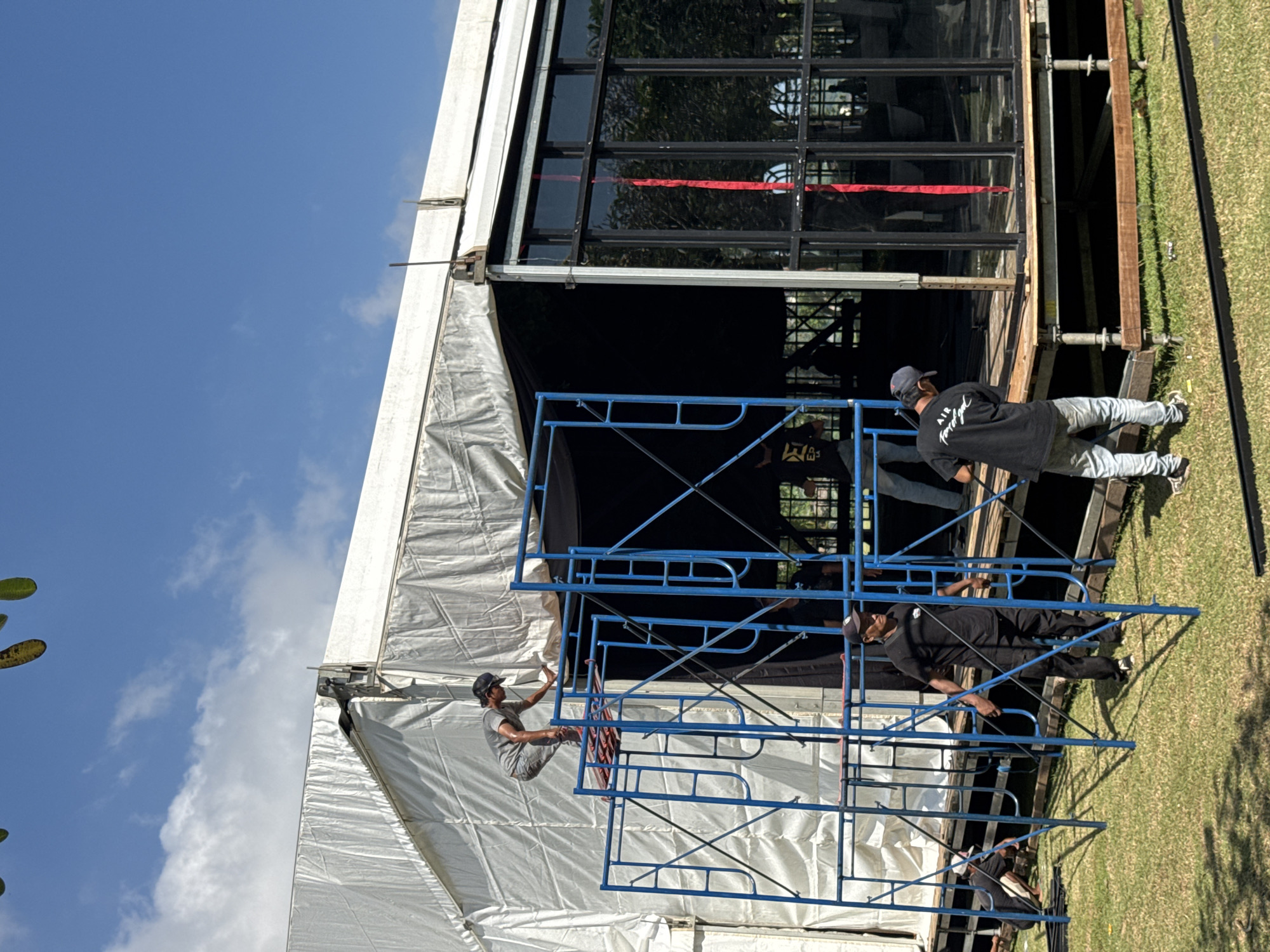 The image shows three individuals working on the exterior of a building under a blue sky. The location is identified as Kuta Selatan, Indonesia. The building appears to be a large structure with a white tent-like roof section. Adjacent to the tent roof is a structure with black-framed windows. The workers are positioned on a blue scaffolding structure. One individual is seated on the scaffolding closer to the tent, while the other two are standing on the scaffolding near the windowed section of the building. One of the standing individuals is wearing a black shirt and a baseball cap. The environment includes green grass and trees visible outside the building structure.