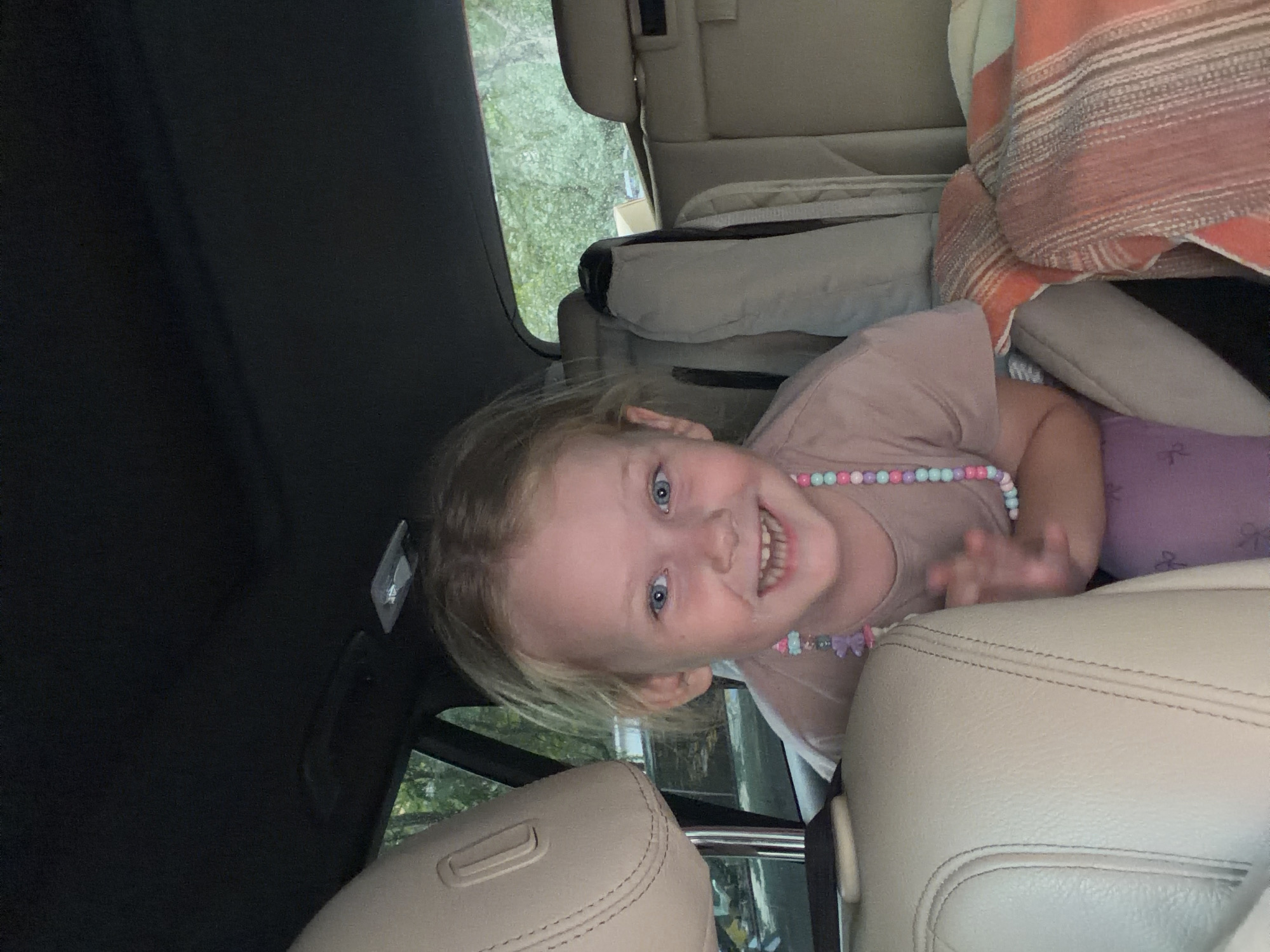 A young girl with blonde hair and blue eyes smiles broadly while looking at the camera from the back seat of a car. She is wearing a light brown t-shirt and a colorful beaded necklace. The car's interior, including beige leather seats and a sunroof, is visible. Outside the car, through the windows, trees and greenery can be seen, suggesting a rural or suburban setting. The image does not provide any information to determine the location as Moscow, Russia.