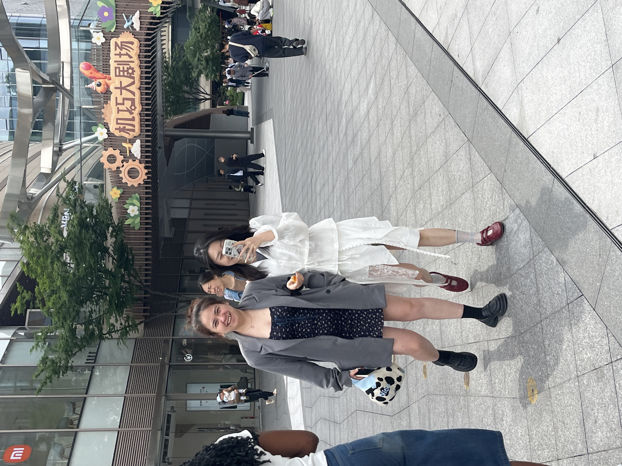 The image shows a group of at least five people outdoors. Two young women are standing in the foreground, smiling. One is wearing a gray blazer over a dark dress with a cow print bag; the other is wearing a white dress and is holding a cell phone as though taking a photo. Behind these women, another young woman is visible. In the background are other pedestrians and a sign written in Chinese. The architectural setting is modern, suggesting an urban commercial area, likely in Liangjiang, China.