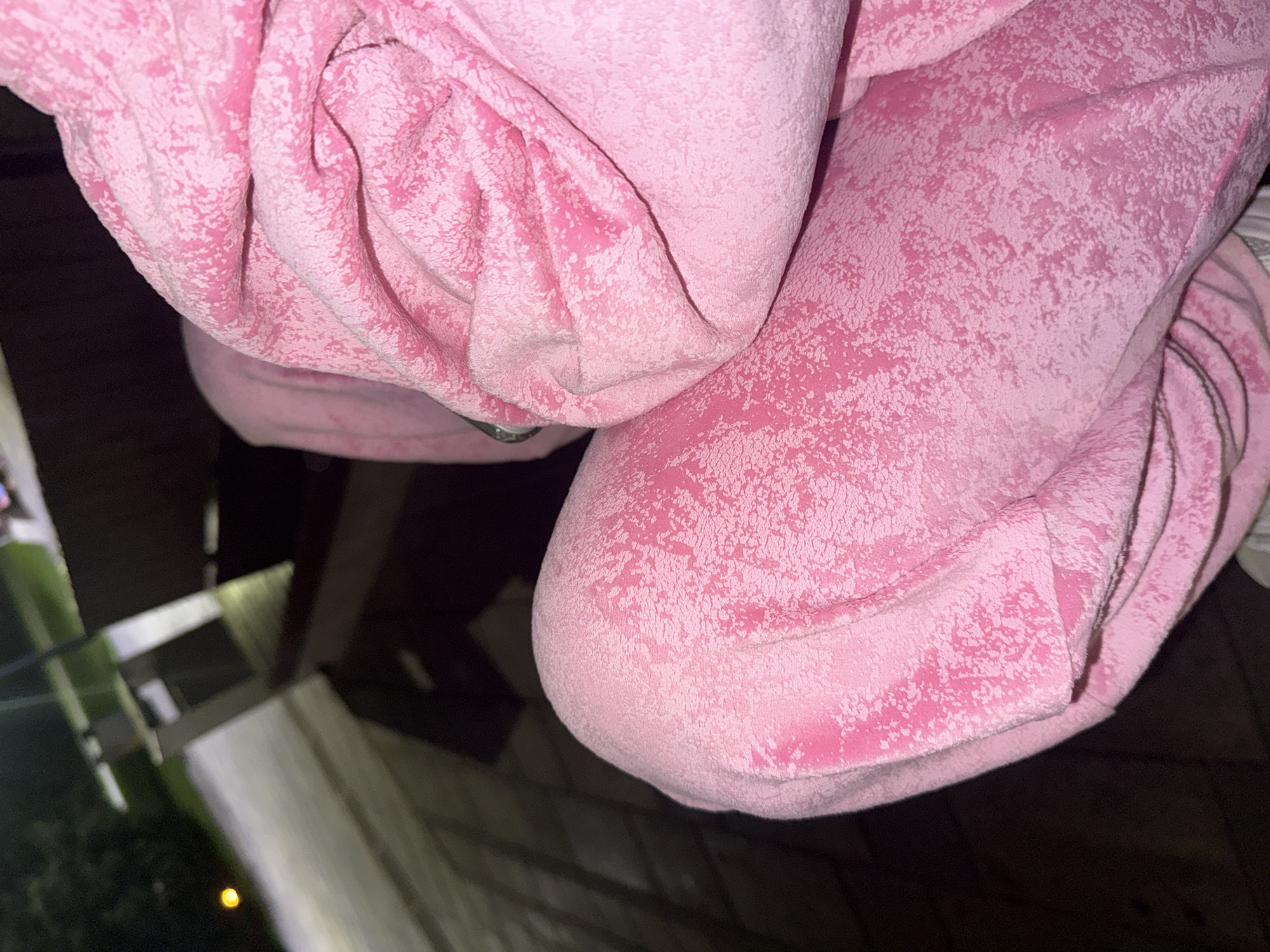 The image shows a close-up view of a person's legs and upper body clad in pink sweatpants and a hoodie. The fabric has a textured, almost speckled appearance with lighter pink and white splotches against a base pink color. The person is likely sitting, as evidenced by the way the fabric folds around their knee and torso. The background is out of focus and appears to be an outdoor setting at night, possibly a park or public area with paved ground and some shadowy structures. There's a hint of greenery and a distant light source on the left side of the image. The location is identified as Dnipro, Ukraine, although the specific context of the scene within the city is not discernible from the image alone.