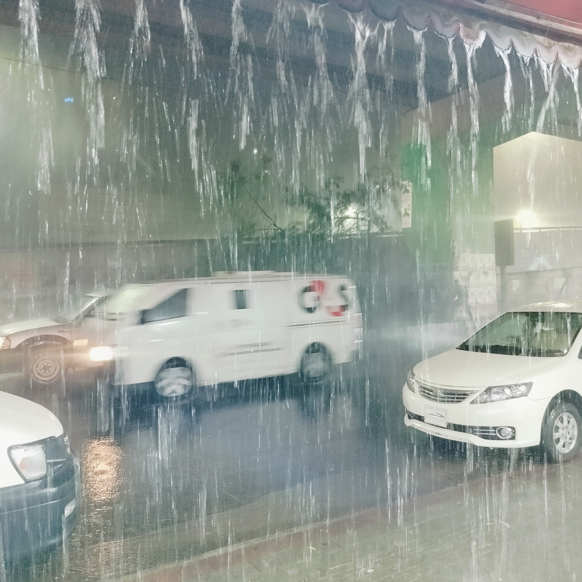 The image captures a heavy downpour on a street in Dhaka, Bangladesh. Several vehicles, including a white van with the letters "GS" on the side and two white cars, are partially visible through the rain. The rain is so intense that it obscures the background, making it difficult to discern specific details of the surroundings. The road is wet and reflects the lights from the vehicles. Above the cars, there's a slight overhang, likely part of a building, from which the rain is cascading.