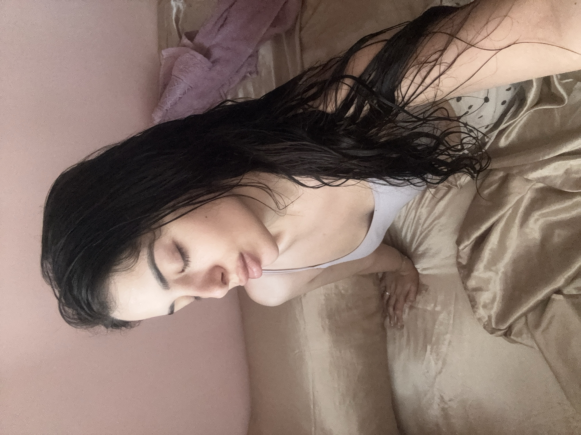 A woman with long, dark, wet hair lies on a golden-colored bed with a pink wall visible in the background. A light purple towel is partially visible on the bed, next to the wall. The woman is wearing a light-colored tank top, and her eyes are closed. Her right hand is visible on the bed, with a ring on one of the fingers. The location is unknown and cannot be determined from the image. The location in Plovdiv, Bulgaria cannot be confirmed.