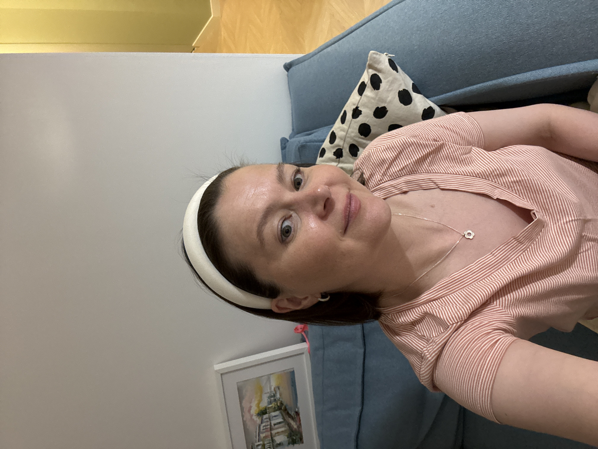A woman with dark hair and a white headband lies on a blue sofa with a black and white spotted pillow. She wears a pink and white striped shirt and a silver necklace with a flower pendant. A framed painting is visible on the wall behind the sofa. The location is not discernible from the image.