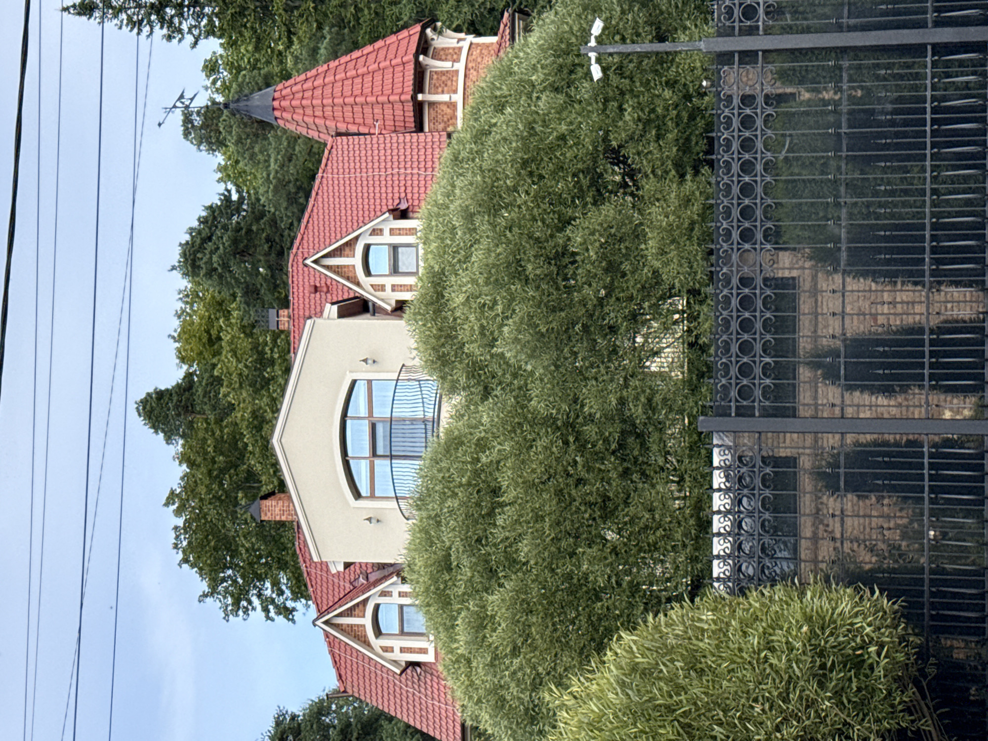 This is a medium-distance, eye-level, outdoor shot taken in Krasnoye Selo, Russia, showing the upper portion of a large, multi-story residence partially obscured by dense, green foliage. The house features a prominent red-tiled roof with dormer windows and a turret with a pointed roof and a weather vane. The building's facade is a mix of light-colored stucco and brick. A black, ornate metal fence runs across the foreground. A street light with two security cameras is mounted on a pole to the right of the frame. The sky is overcast with visible power lines.